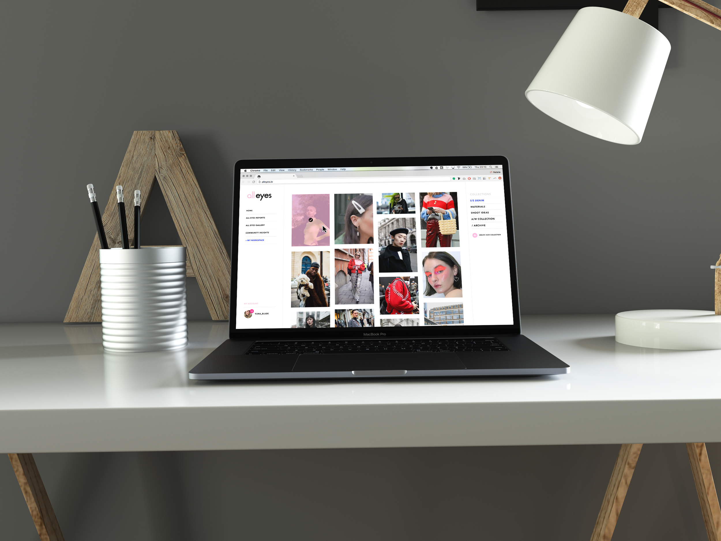 A desk with a MacBook Pro laptop displaying a fashion website, a white cylindrical pen holder with three black pencils, a wooden shelf with a white lamp hanging above, and a wooden support leg visible on the left side.