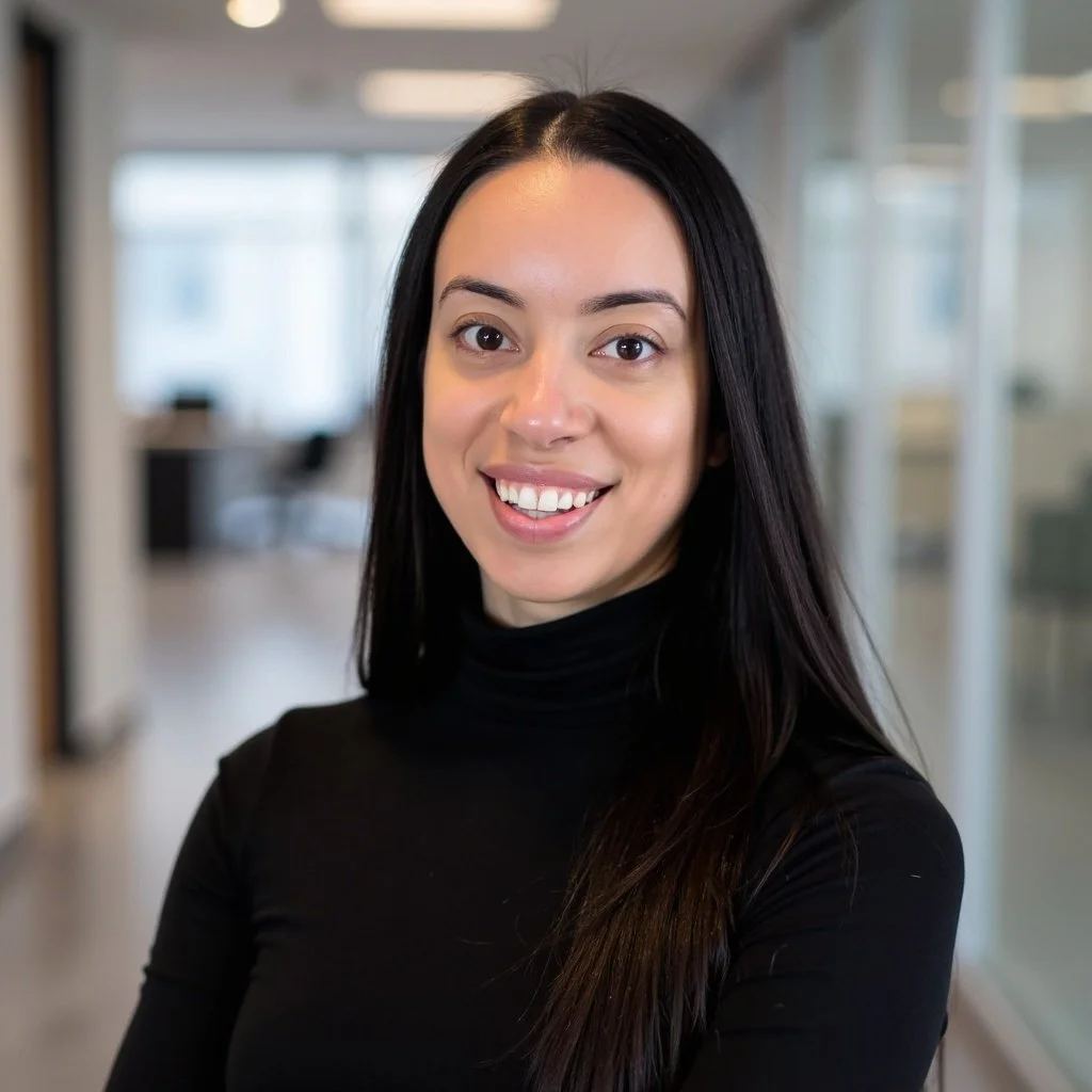 A woman with long dark hair wearing a black turtleneck sweater smiling in a bright office environment.