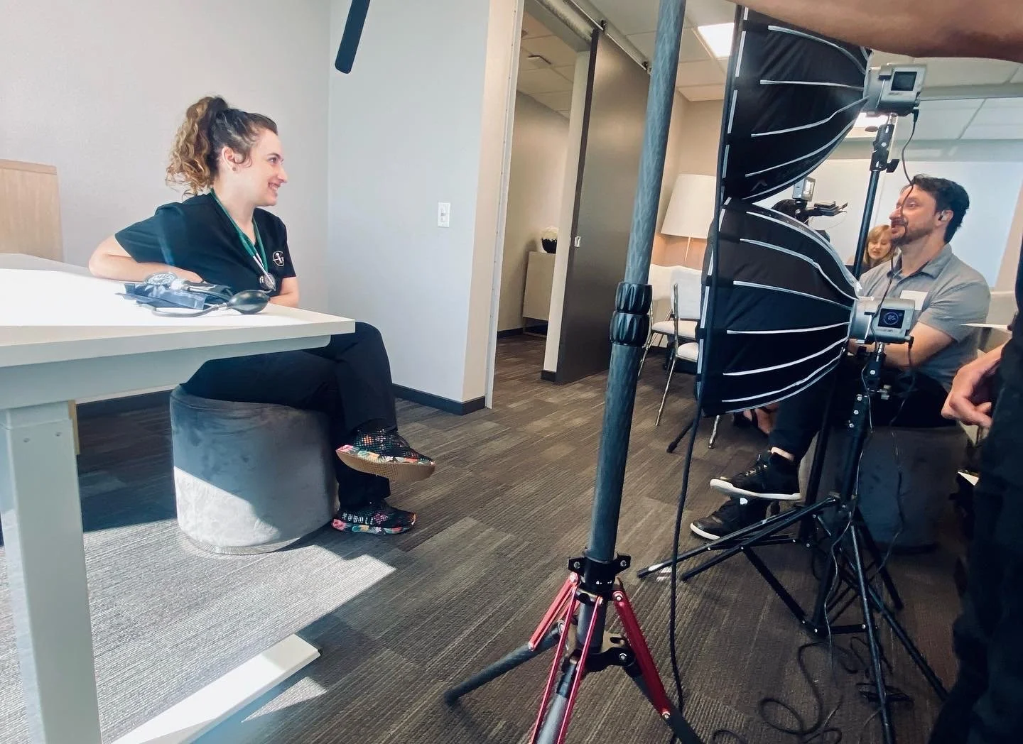 A woman in medical scrubs sitting at a white table during a filmed interview with a man and a person taking notes.