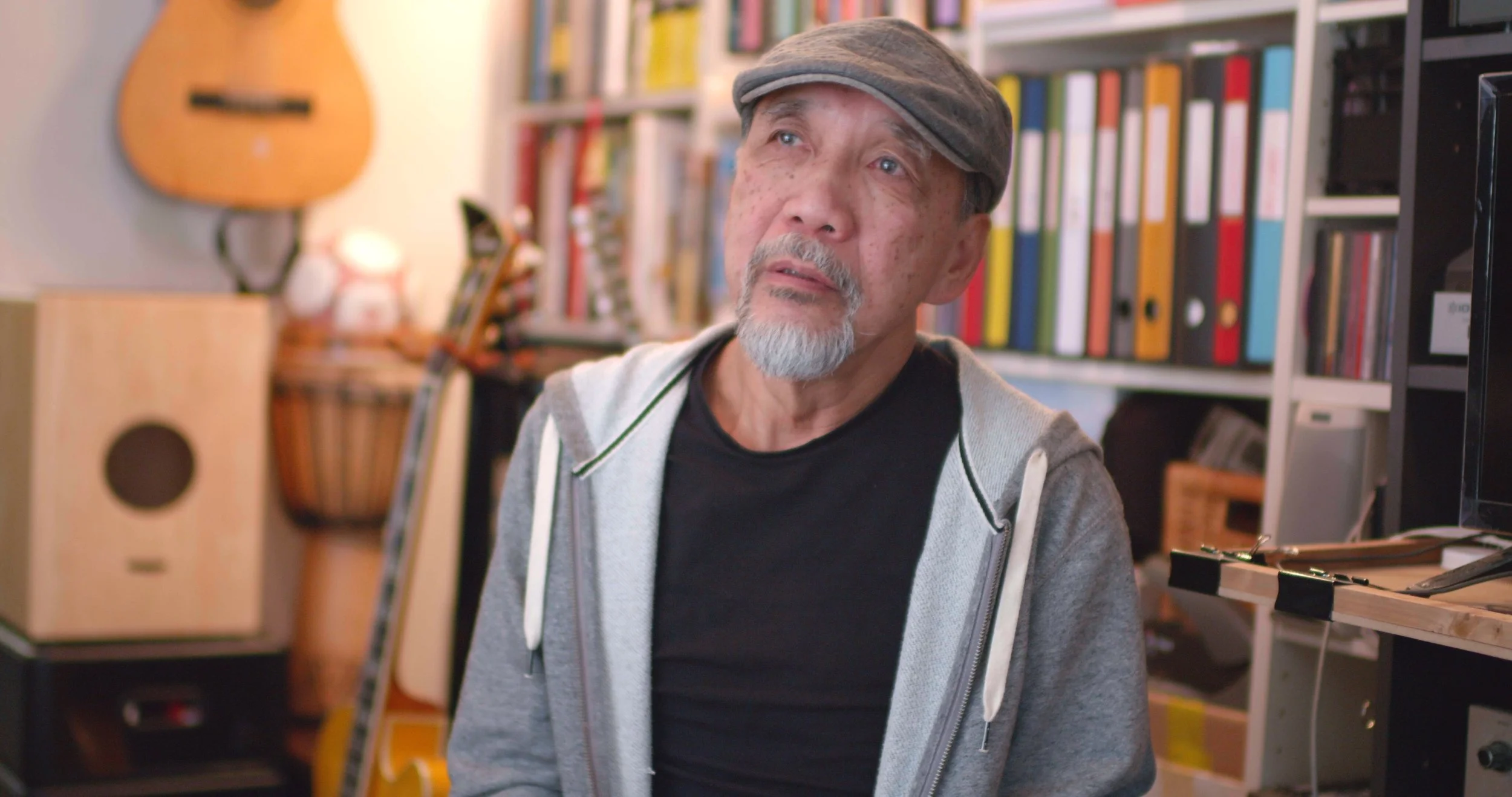 An elderly man with a gray beard, wearing a gray cap and hoodie, sitting in a room with shelves of books and musical instruments, looking up thoughtfully.