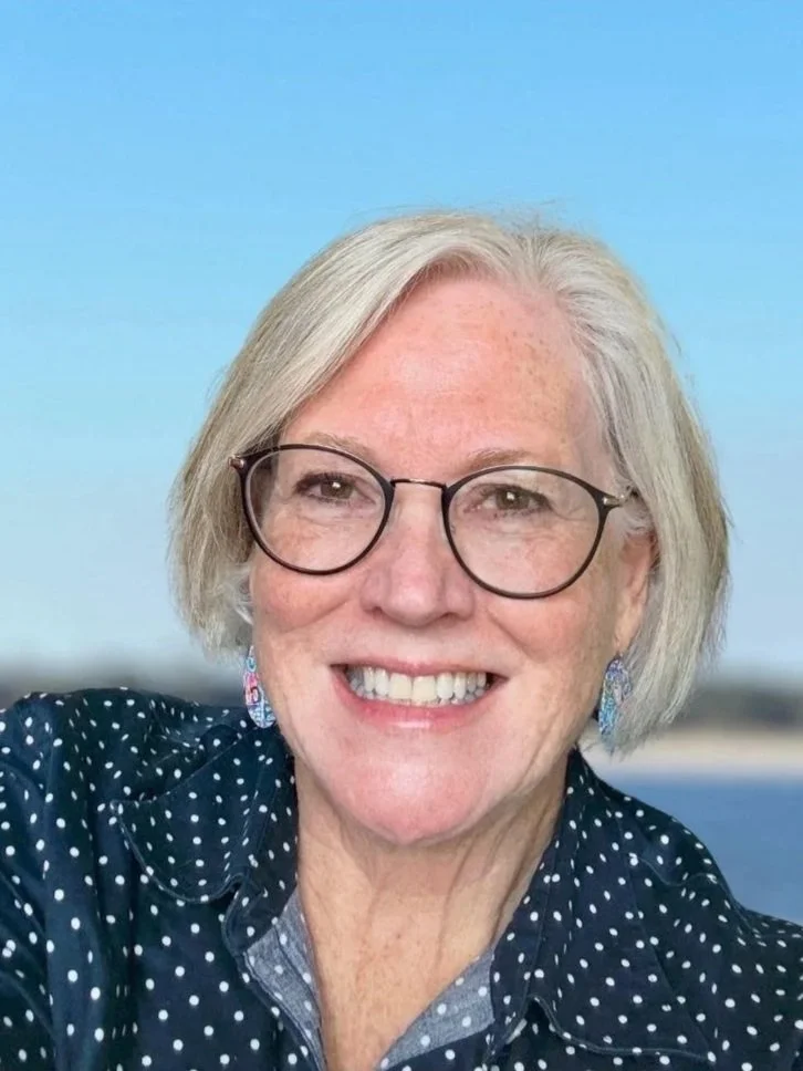 A smiling woman with short gray hair, glasses, earrings, and a dark blue polka dot shirt against a blue sky background.