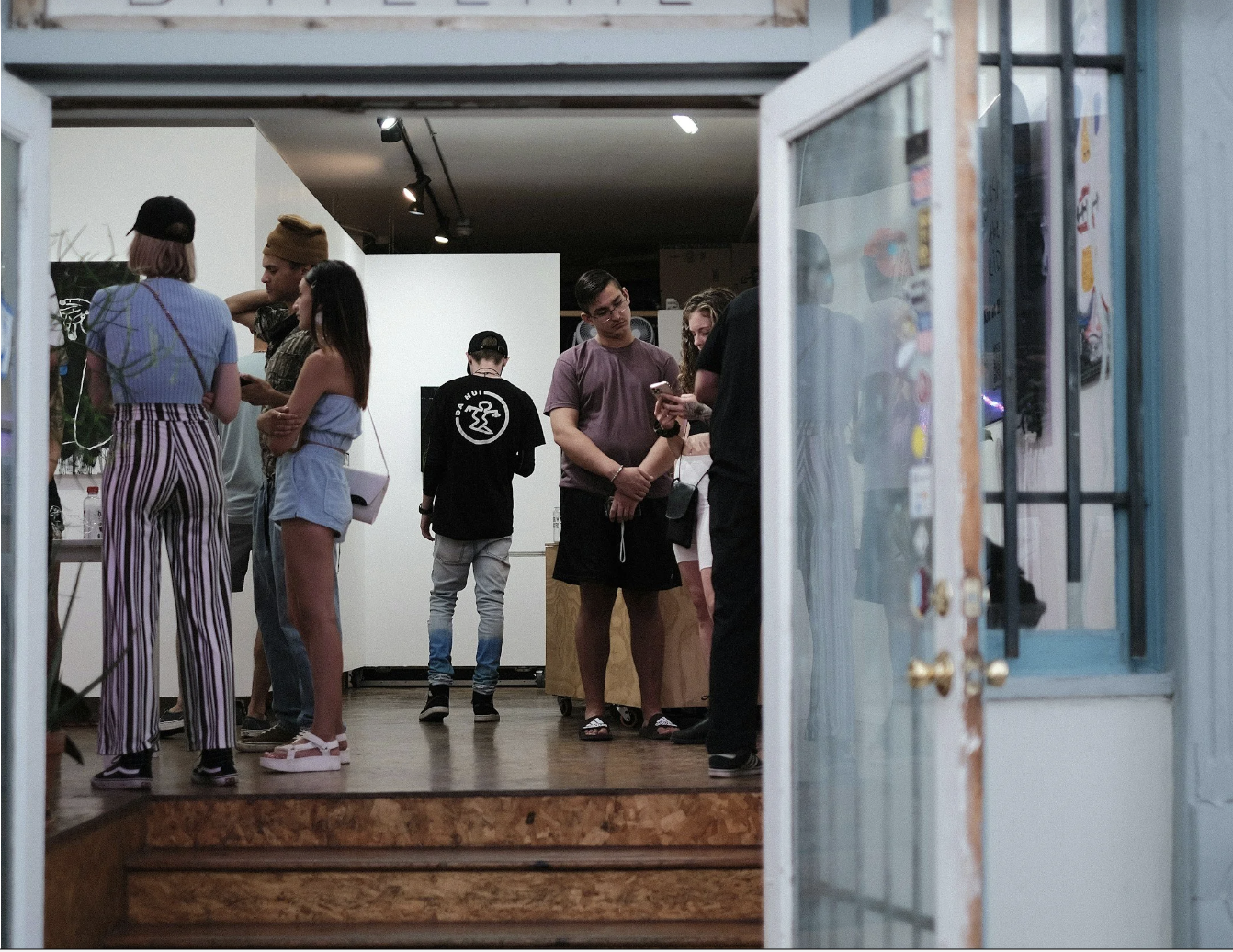 People gathered in an art gallery, with some looking at their phones and others talking, seen through an open door.
