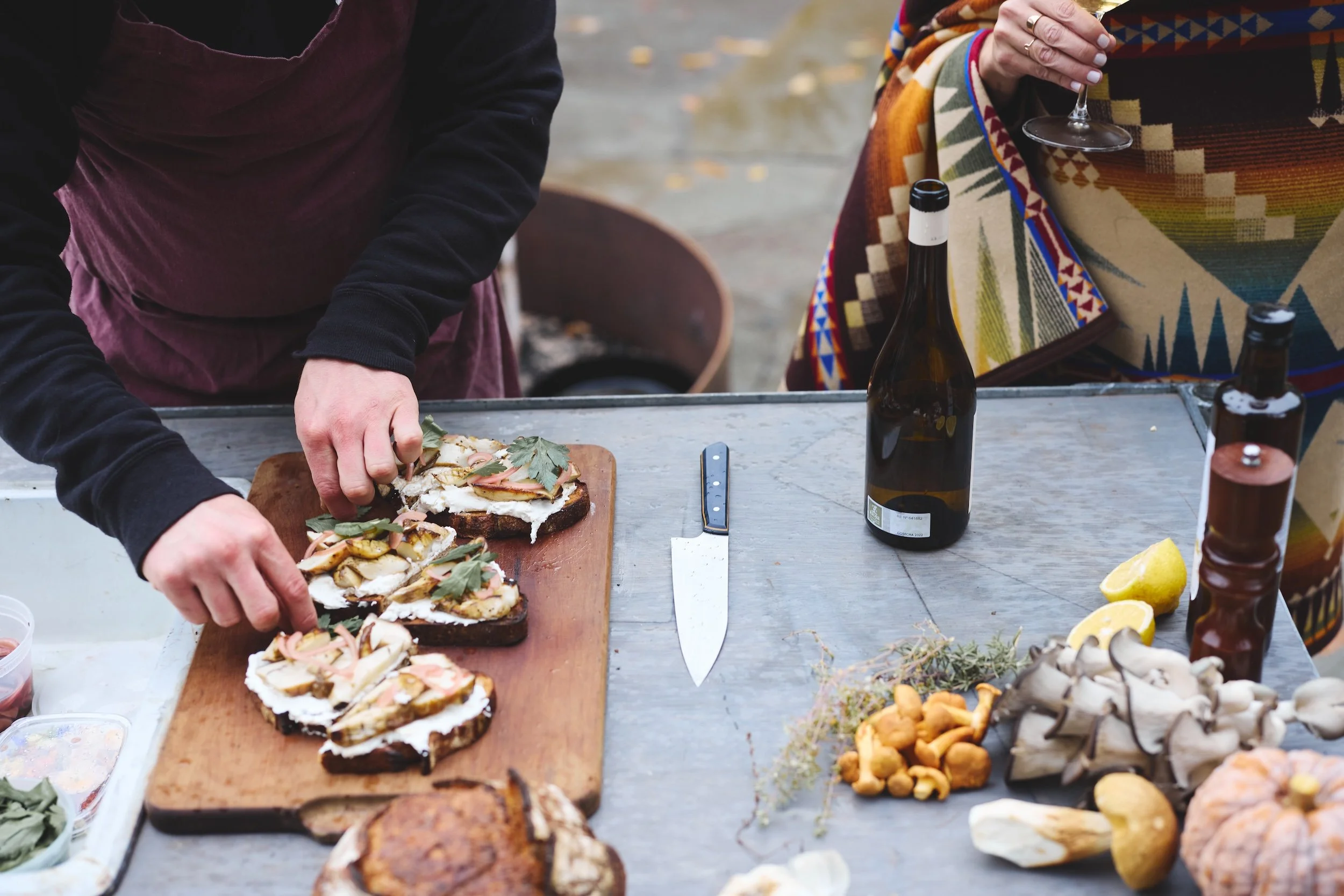 Person preparing sandwiches on a wooden cutting board outdoors, with bottles of beer, lemon wedges, and food items on the table.