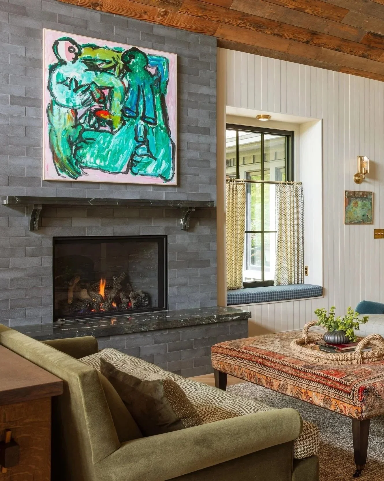 Living room with a gray brick fireplace and a colorful abstract painting above it. A window with yellow patterned curtains, a cushioned window seat, and a dining table with a decorative tray and plant. Part of an upholstered couch and armchair are also visible.
