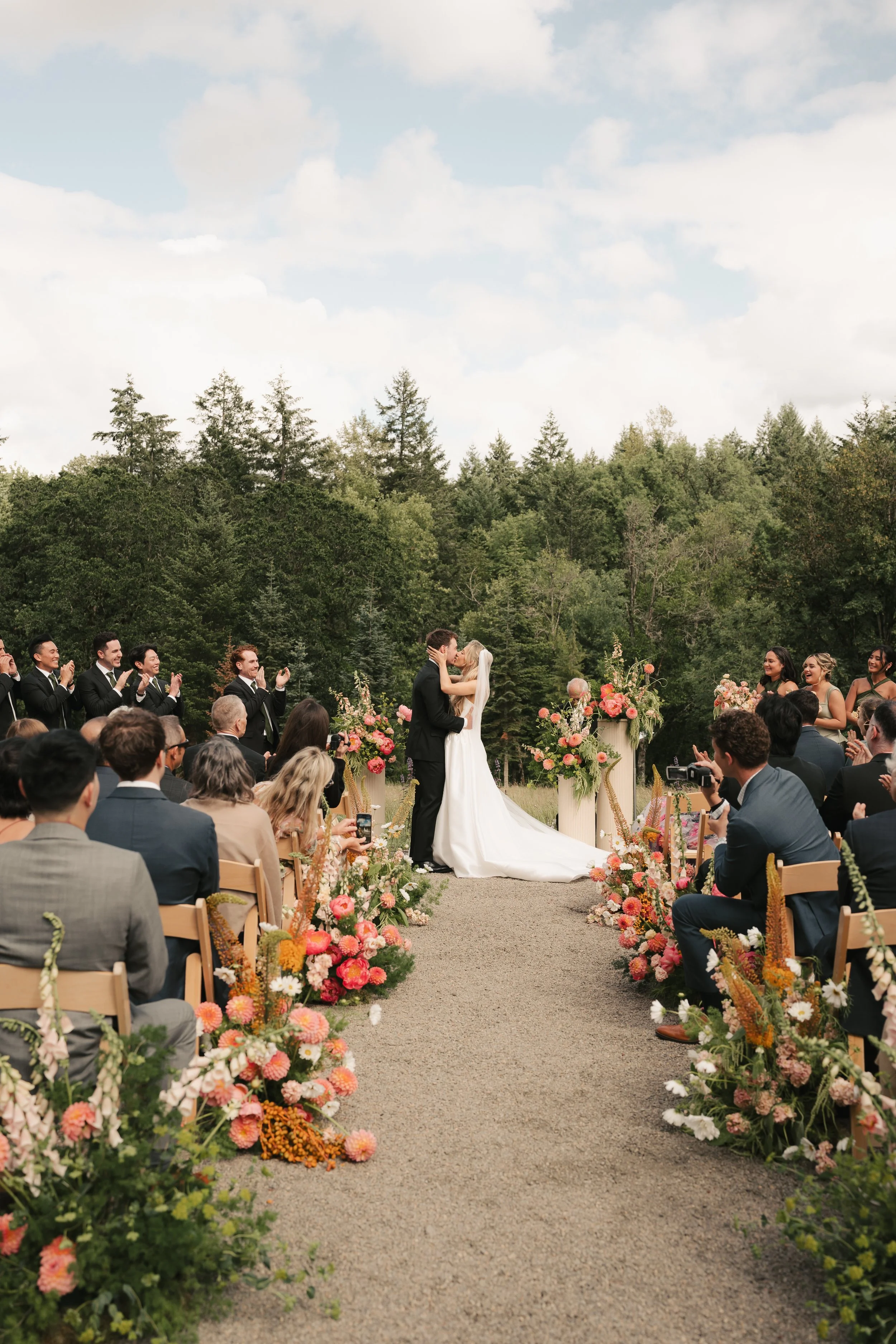 A wedding ceremony outdoors with a bride and groom kissing, surrounded by friends and family, with colorful flower arrangements and a backdrop of forest and sky.