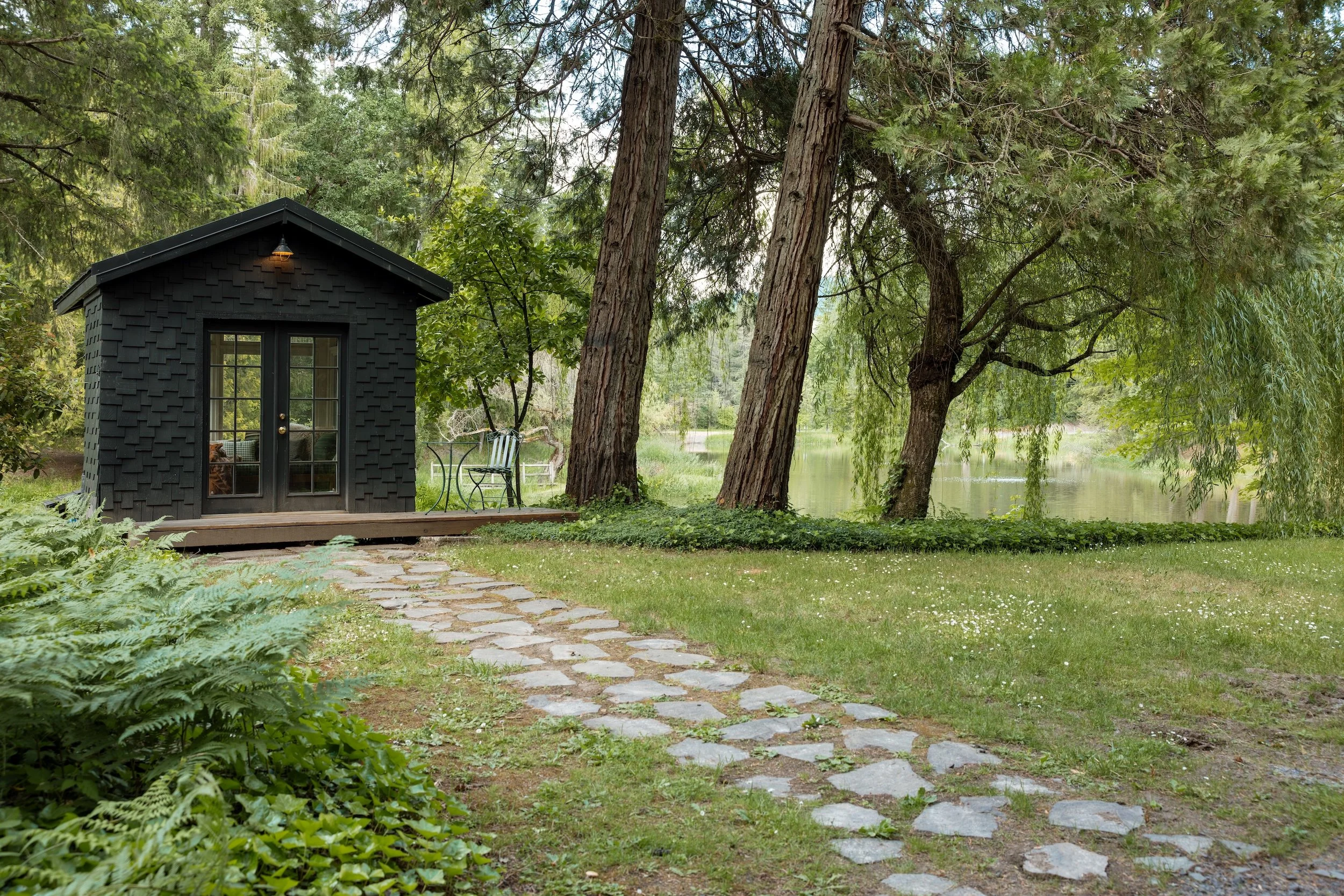 A small black shed with a door and window, surrounded by green trees and grass, with a stone pathway leading to it by a lake or pond.