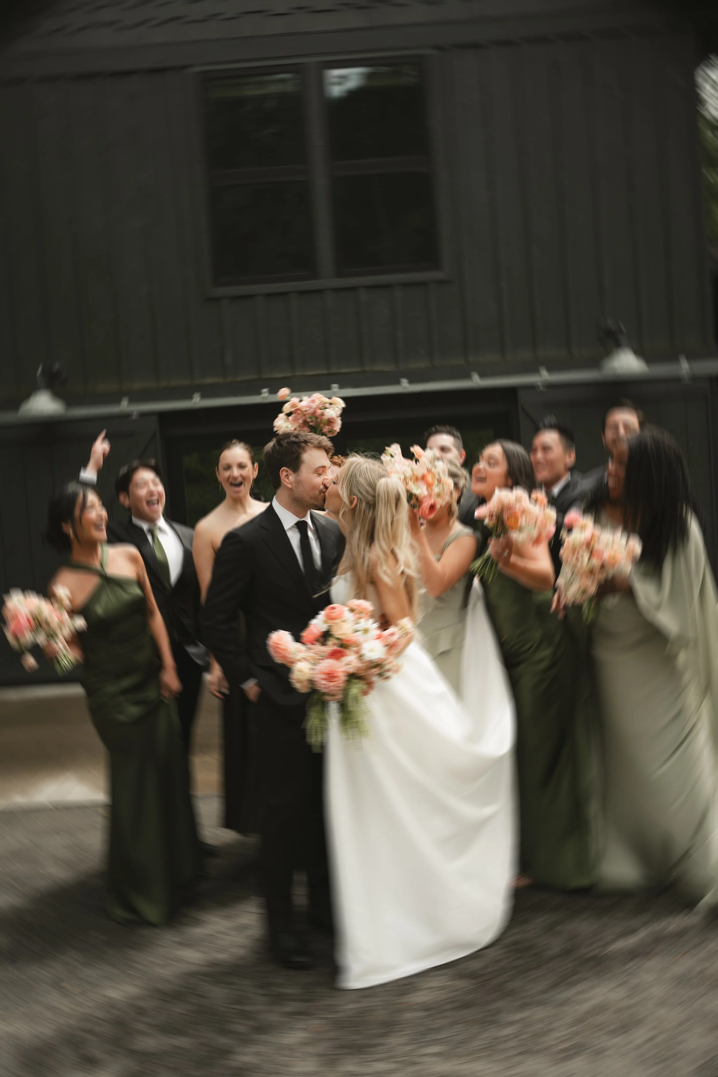 A wedding celebration with a bride and groom kissing, surrounded by wedding party members holding bouquets, cheering, and smiling outside a building with black siding.