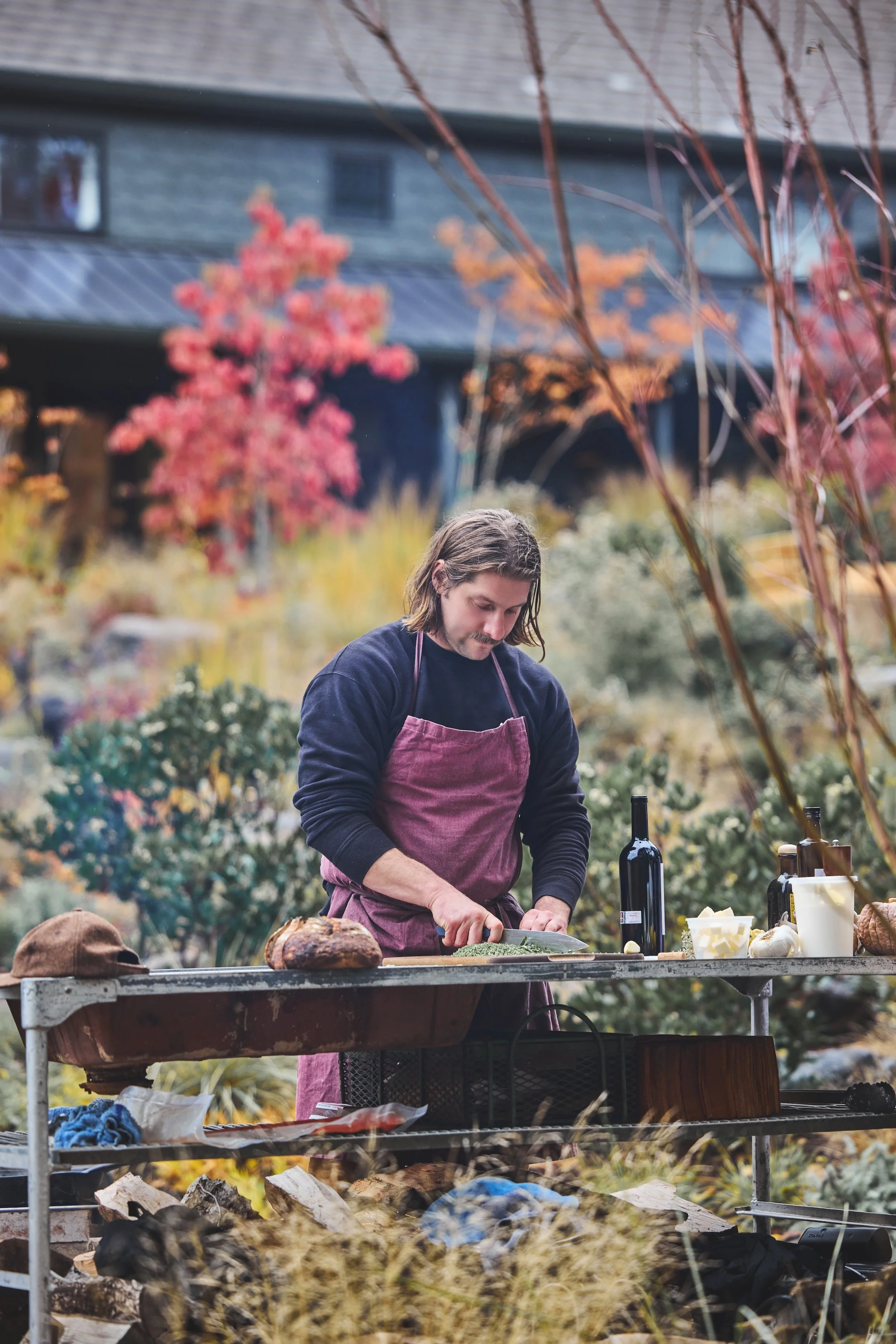A man with long hair and a mustache wearing a pink apron and navy sweatshirt, preparing food outdoors at a table surrounded by plants and trees with red and orange autumn leaves, under a cloudy sky.