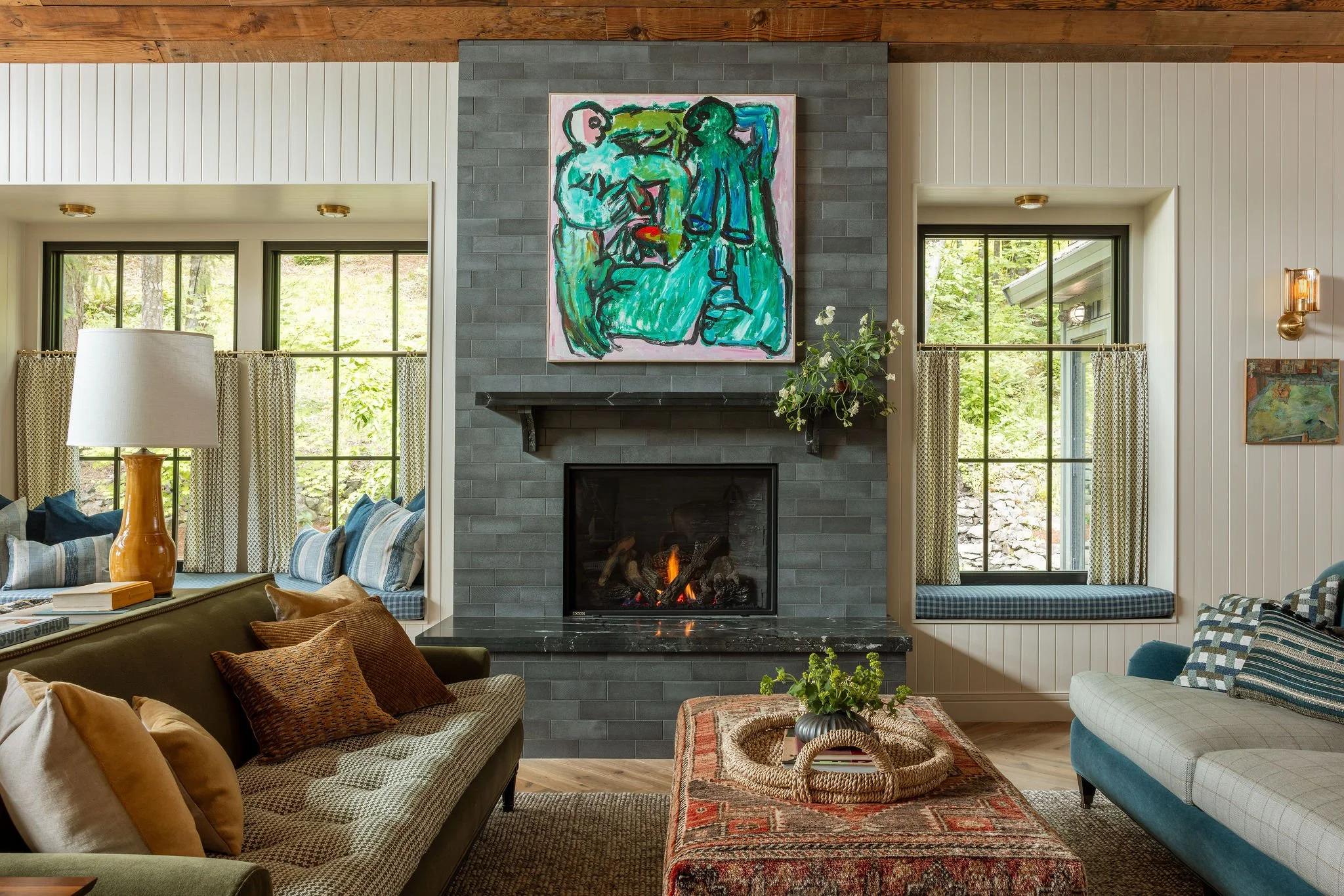Living room with a gray brick fireplace, colorful abstract painting, and large windows showing trees outside.