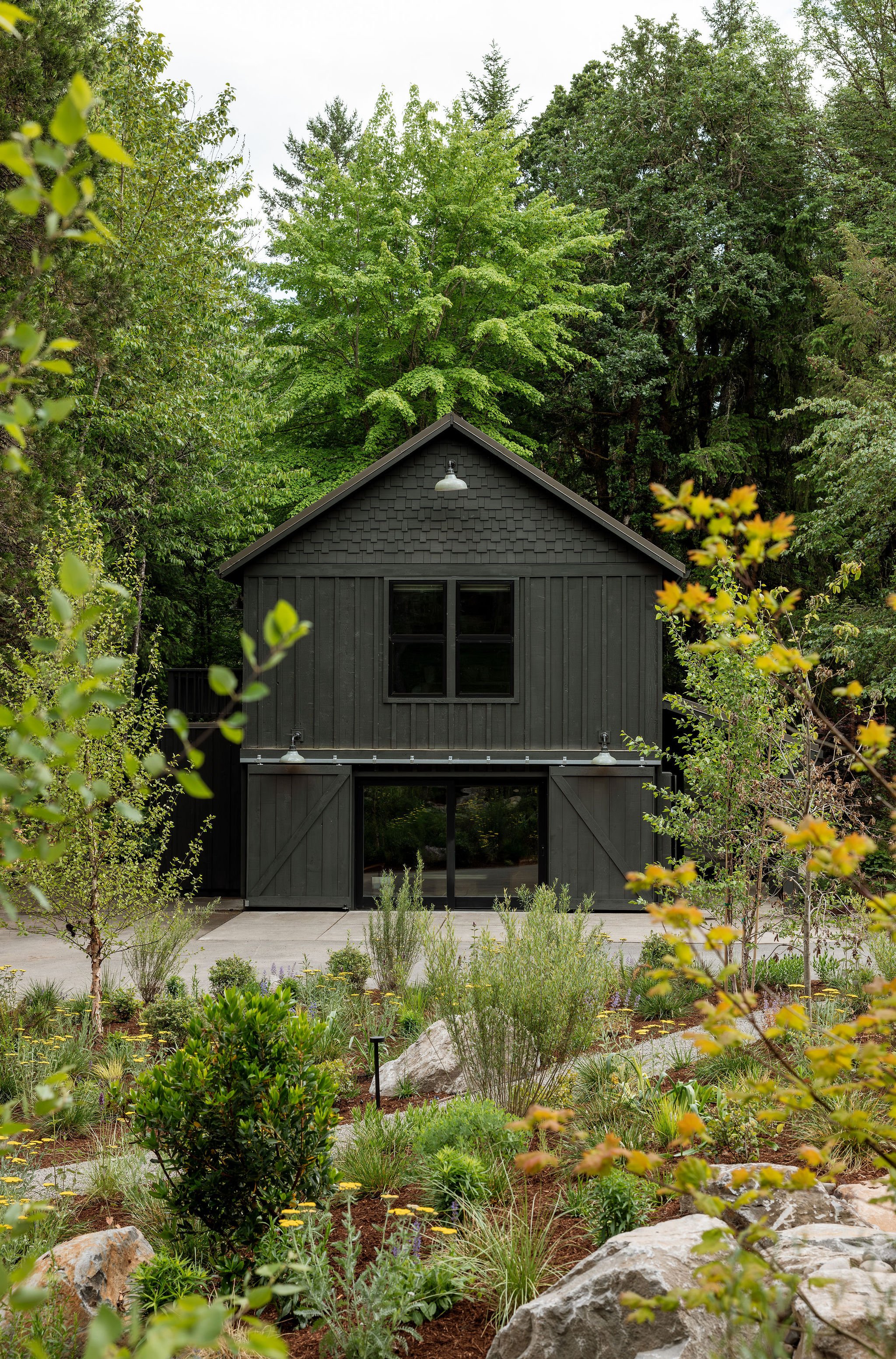 Black barn-style house with large sliding glass door and window, surrounded by green trees and native plants with rocks and mulch in the garden.