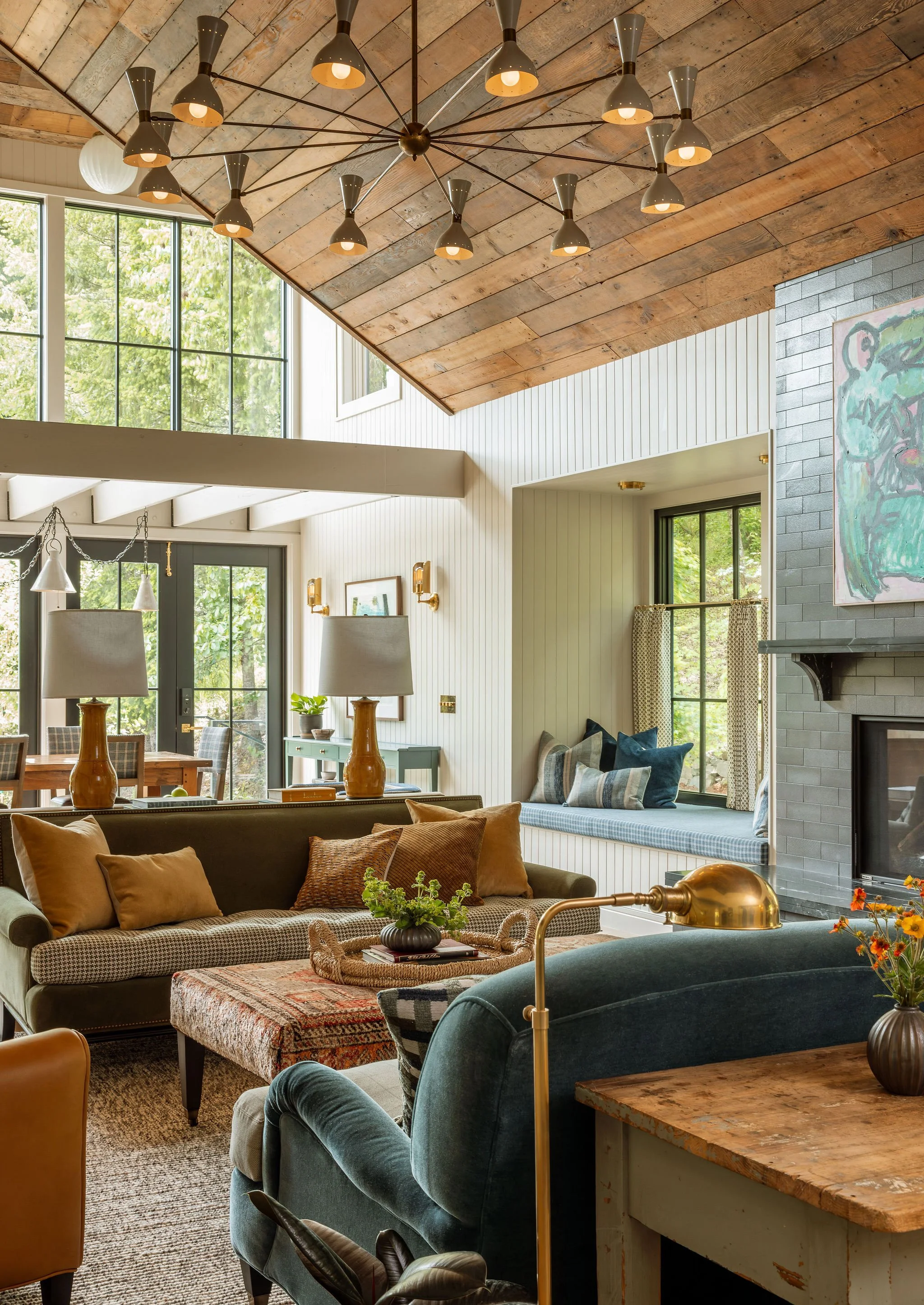 Interior of a living room with a vaulted wooden ceiling, large windows, coffee table, various couches and armchairs, and a fireplace. Natural light illuminates the space.