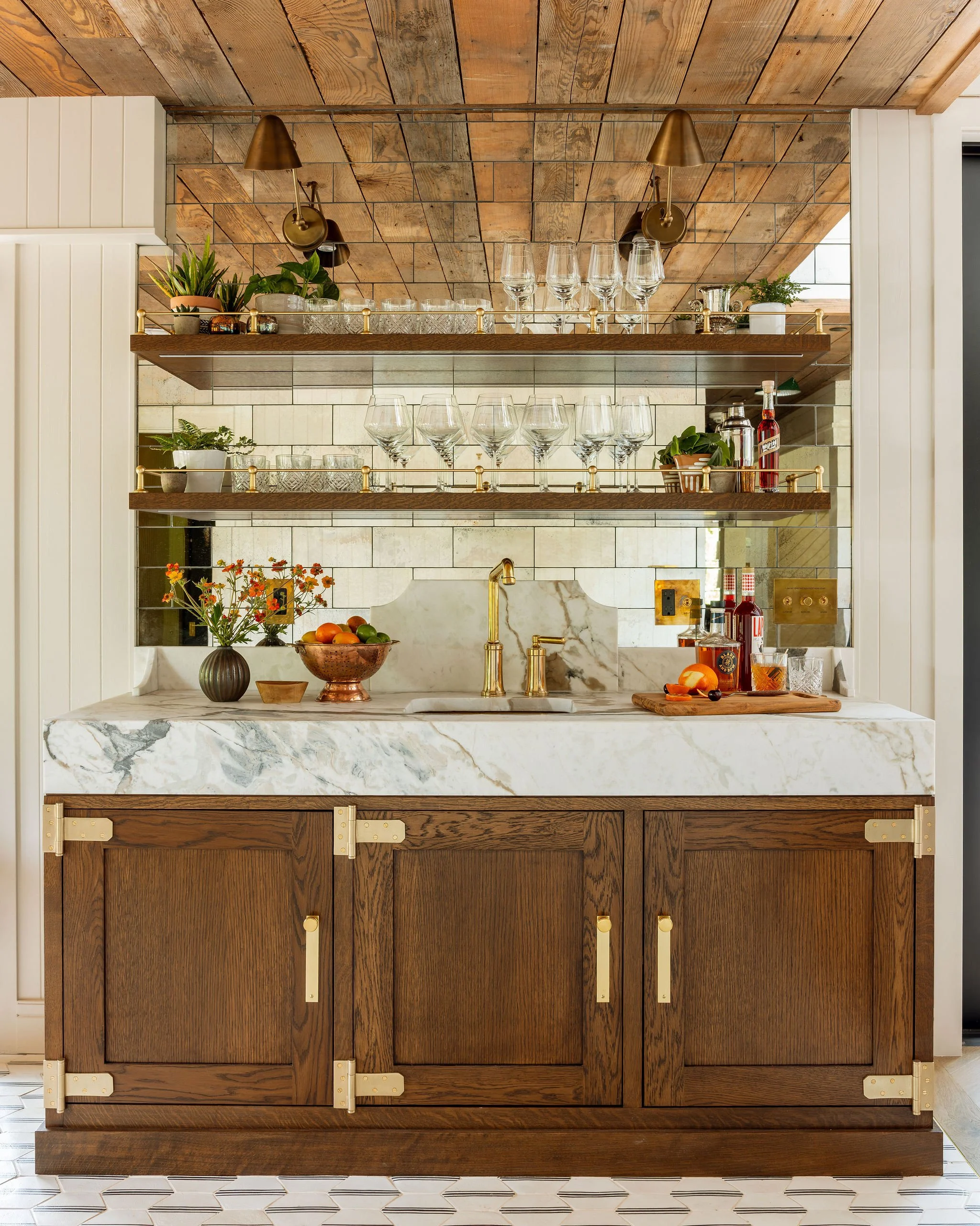 Kitchen with marble countertop, wooden cabinet doors with gold hardware, open shelves with plants, glassware, and bottles, tiled backsplash, gold faucet.