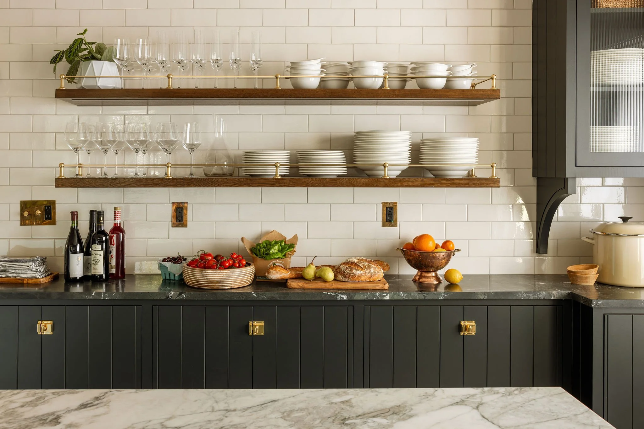 Kitchen shelves with glassware, plates, bowls, and a plant, set against white subway tile backsplash, with fruit, bread, and bottles on the counter.