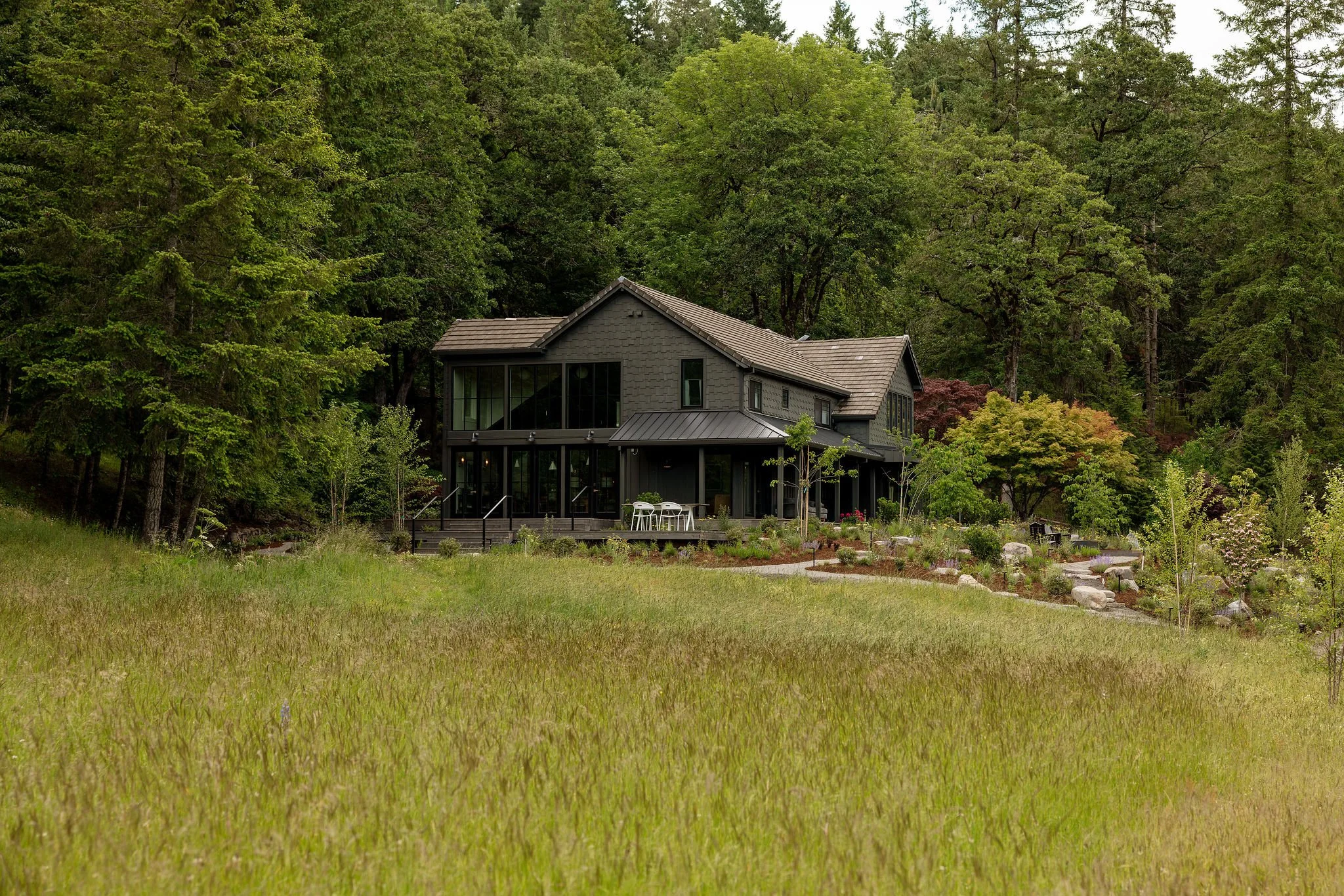 A modern gray house with large windows, surrounded by lush trees and greenery, with a patio and garden in the foreground.