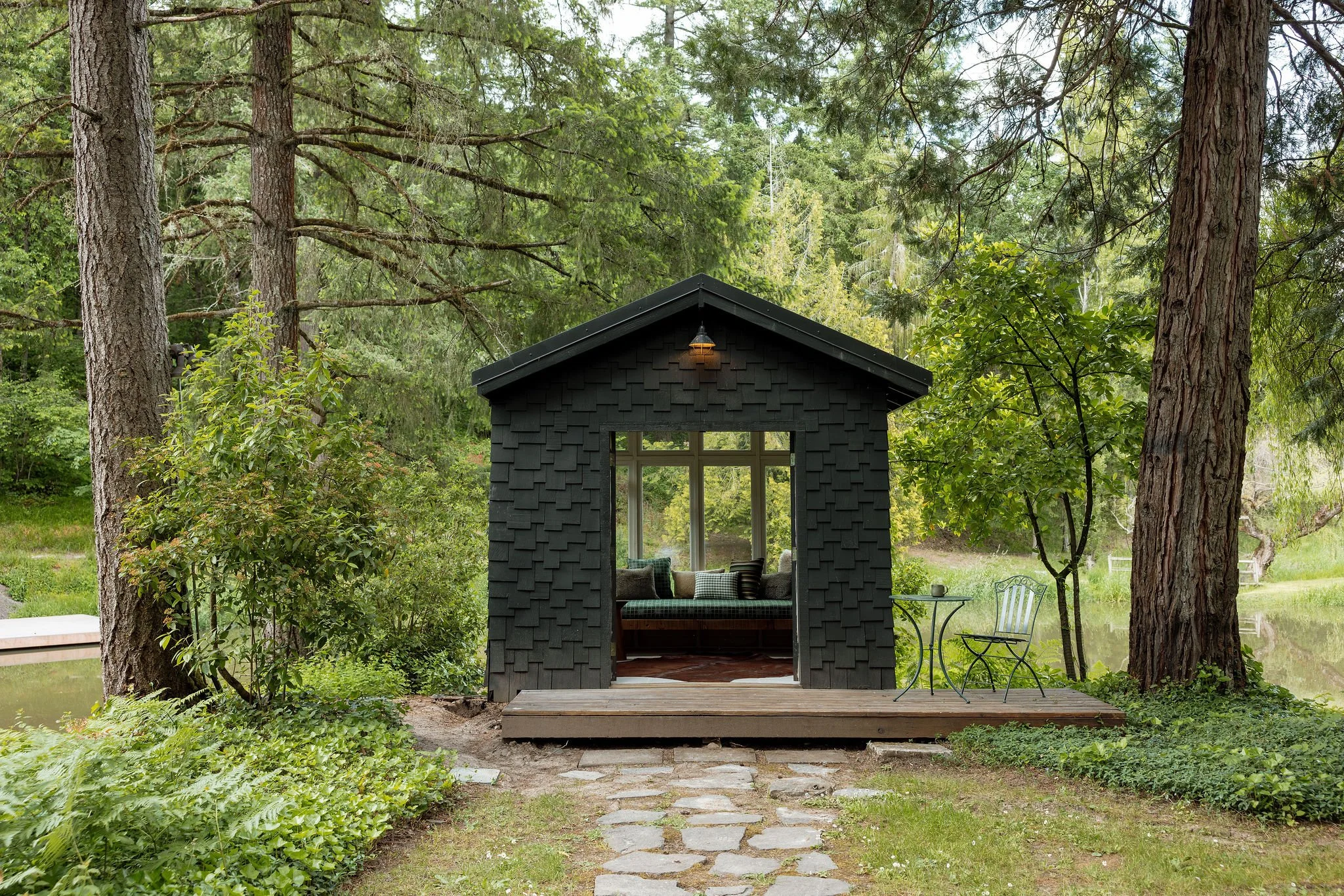 A small black outdoor cabana with an open front and a built-in window seating area, set in a lush green forest with tall trees and a grassy backyard. The cabana has a sloped roof with a light hanging above the window.