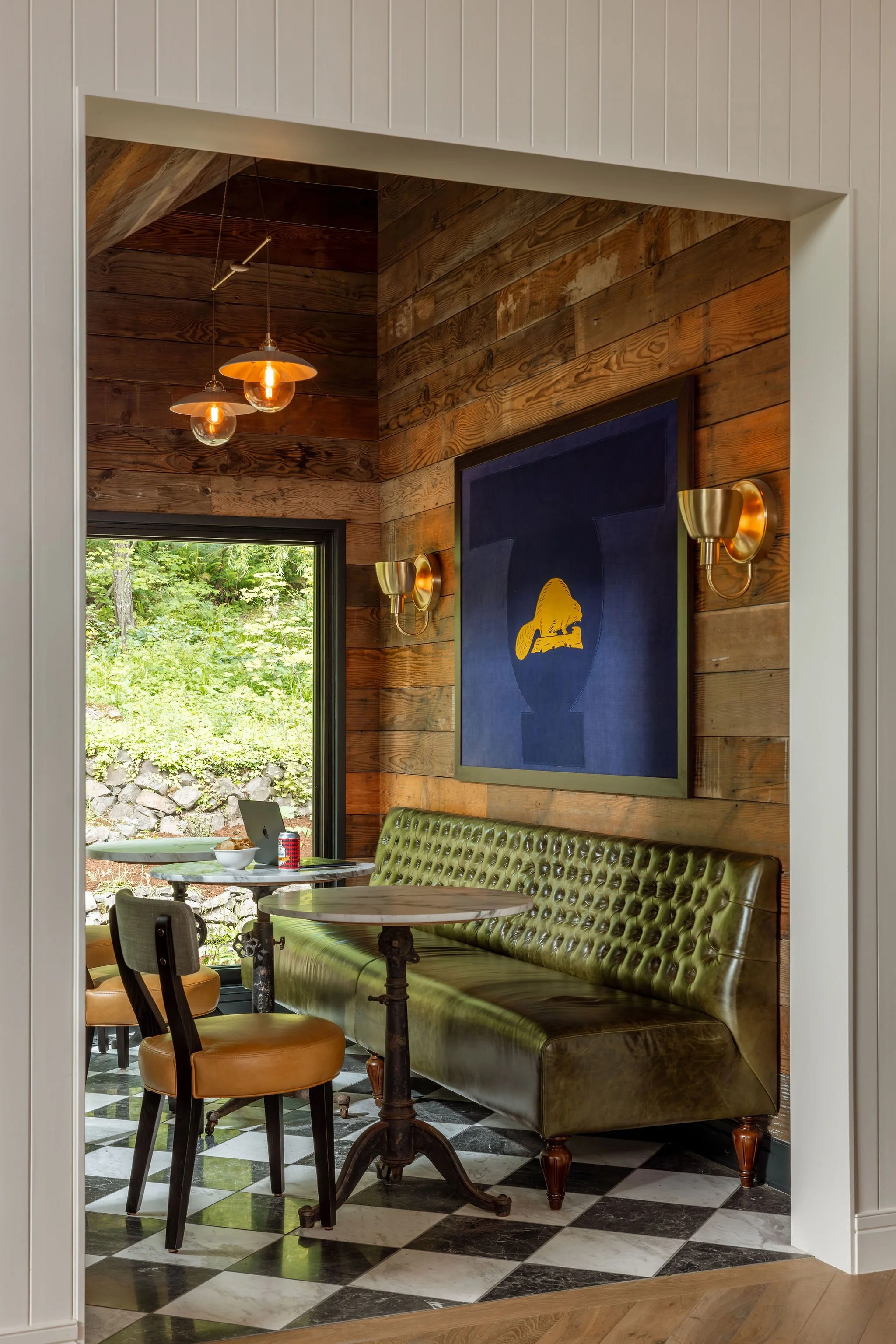 Interior of a cozy room with wood-paneled walls, a window showing greenery outside, a green upholstered bench, a small round table with chairs, and decorative lighting fixtures.