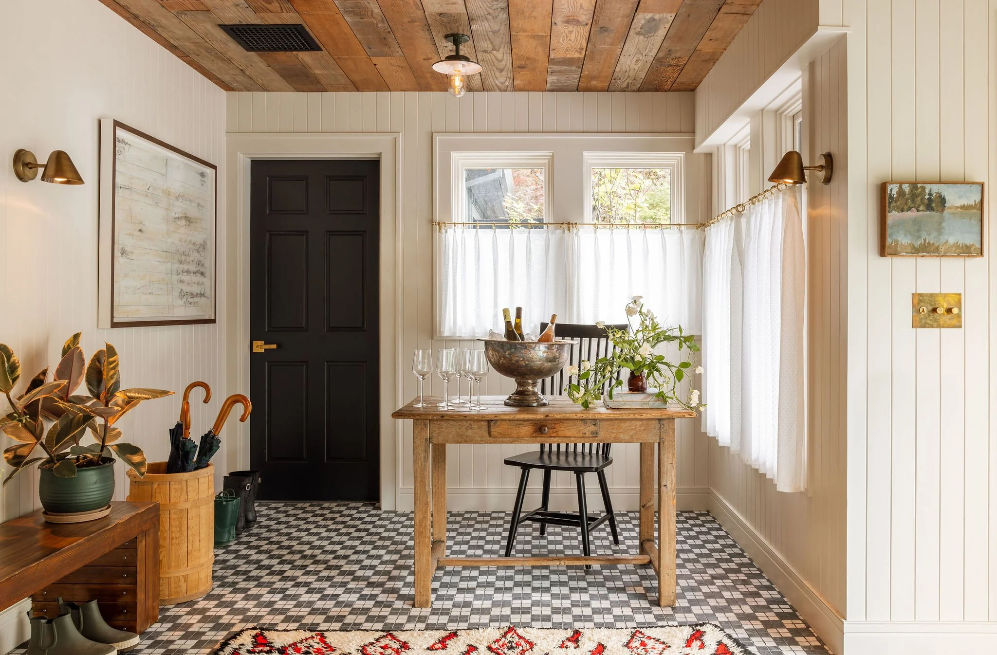 View of a cozy dining area with a wooden table, black chairs, and a bowl of bottles on top, near a window with white curtains. The room features white paneled walls, a dark door, a wooden ceiling, and patterned tile flooring. Plants and artwork are a