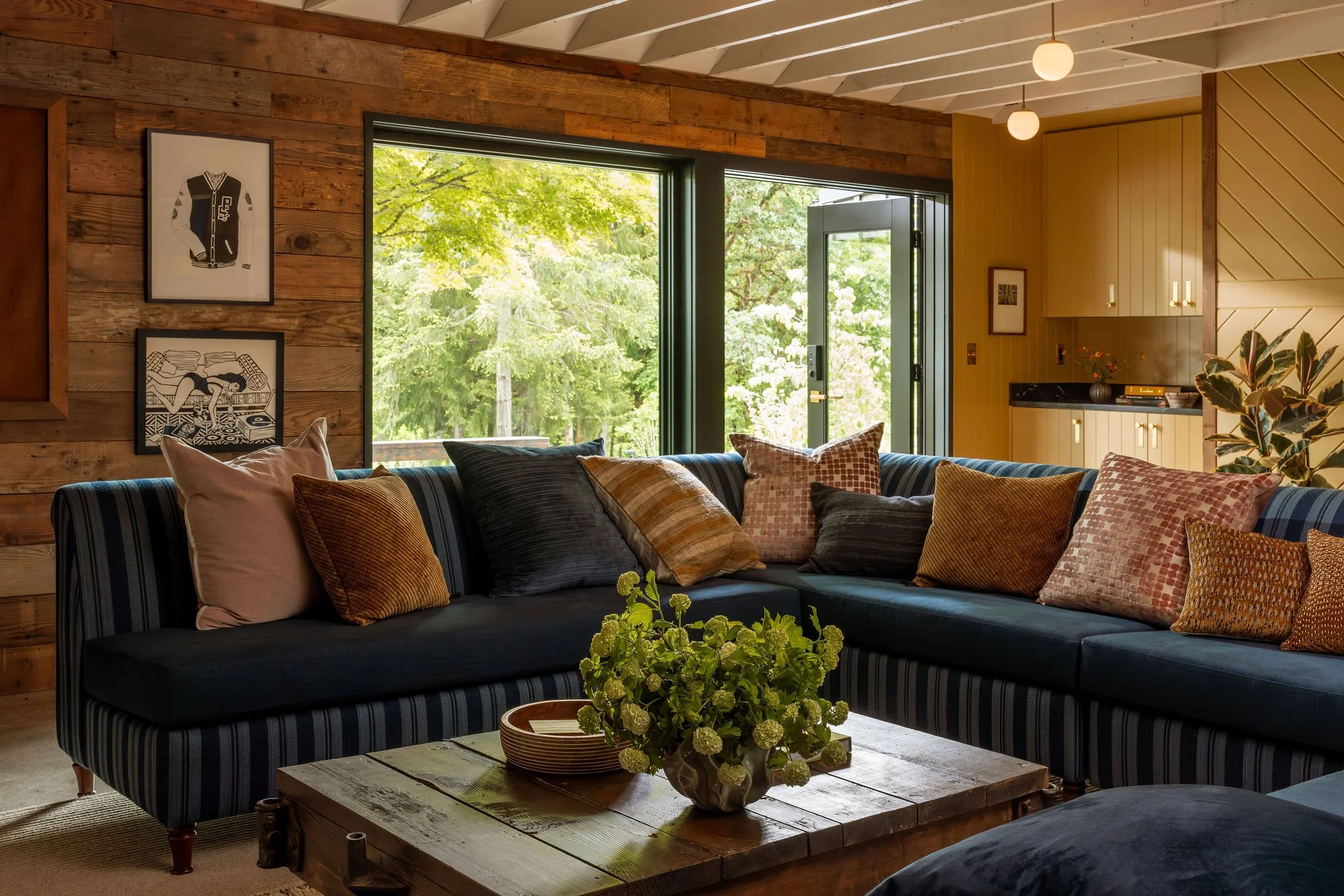 A cozy living room with a dark striped sofa filled with various pillows, a rustic wooden coffee table with a bouquet of green flowers, large glass windows showing lush green trees outside, wooden paneled and yellow walls, framed artwork, and warm lig