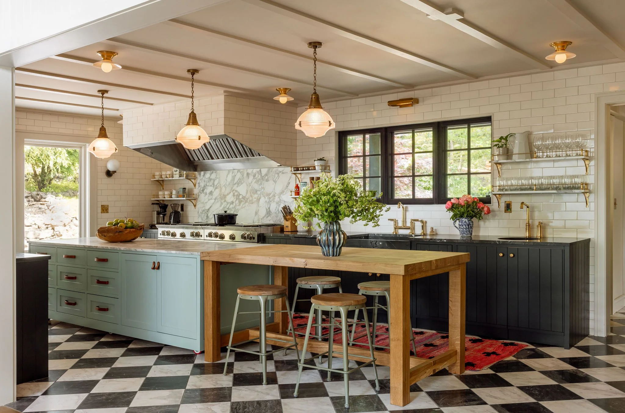 Modern kitchen with black and white checkered tile floor, turquoise and navy blue cabinetry, a white marble backsplash, and gold fixtures. There are pendant lights hanging from the ceiling, a large window with a view of greenery, and decorative items like flowers and glassware on open shelves.