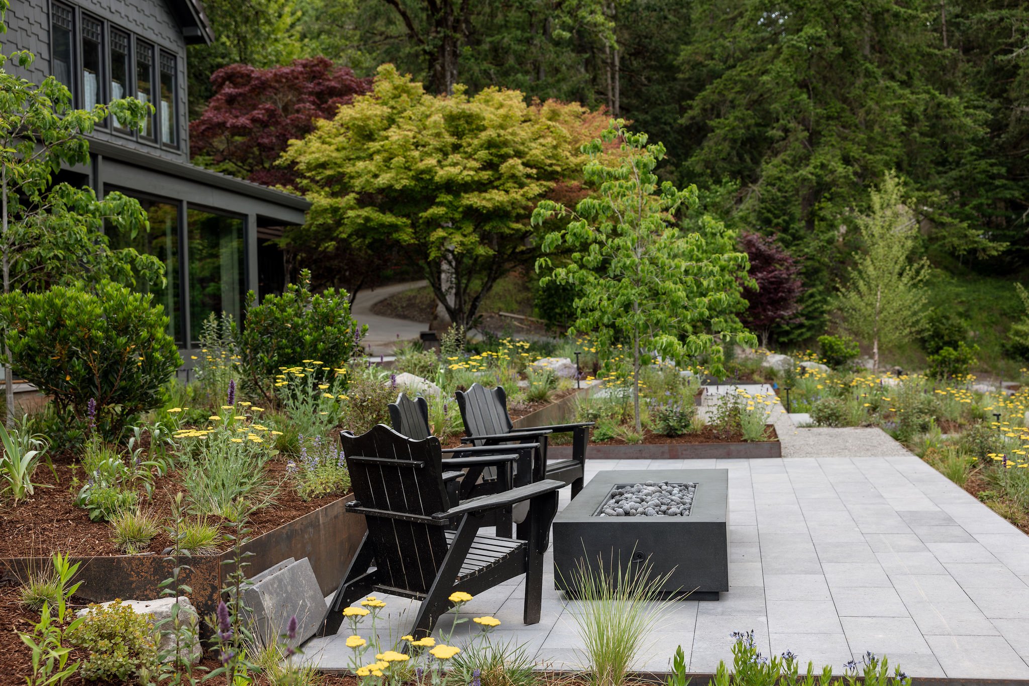 Outdoor garden seating area with black chairs arranged around a fire pit filled with stones, surrounded by lush green trees and flowering plants, with a modern house on the left in the background.