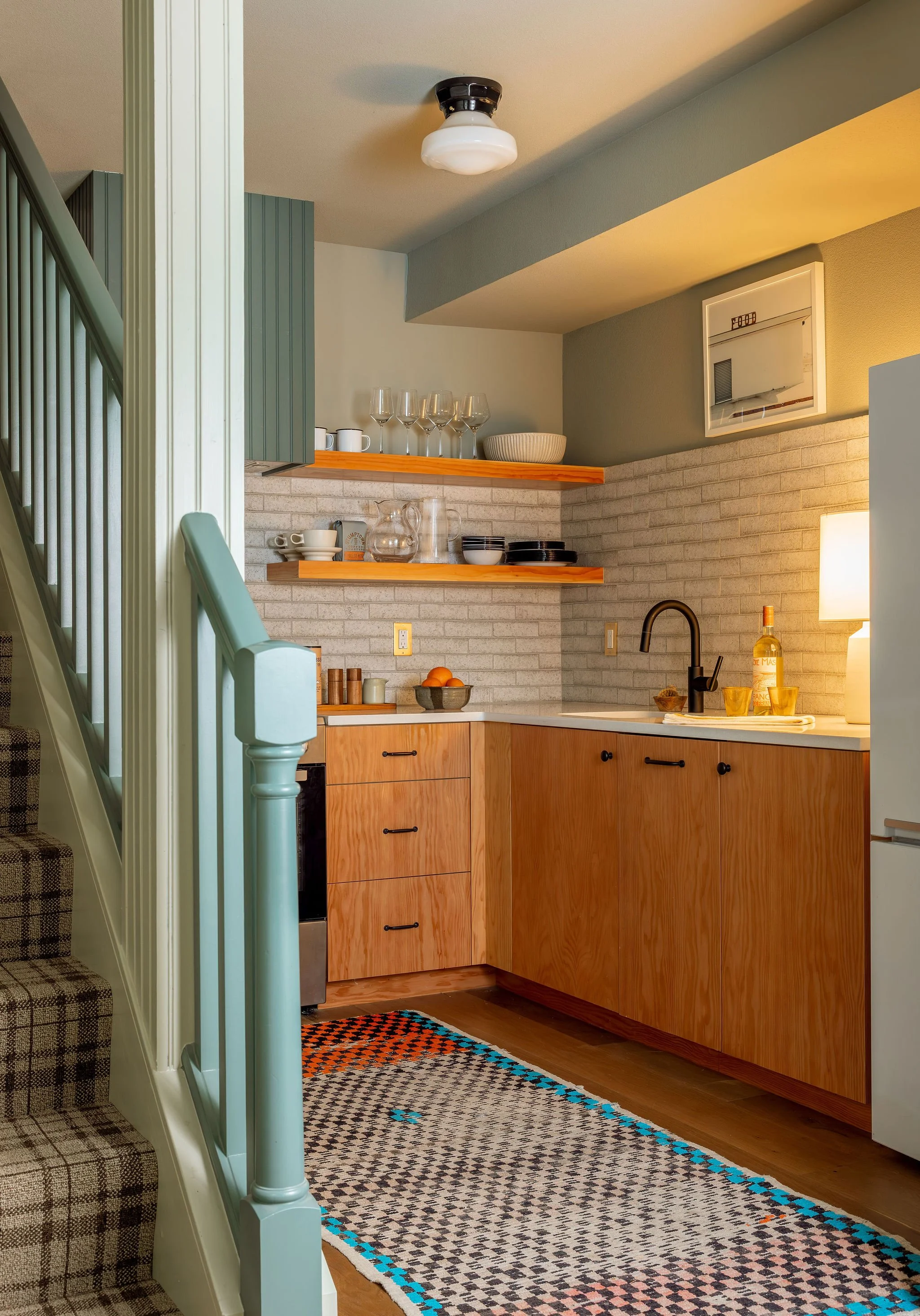 A cozy kitchen area with wooden cabinets, open shelves holding glassware and dishes, a black faucet at the sink, and a patterned rug on the hardwood floor. An adjacent staircase with a pastel mint green handrail is visible on the left.