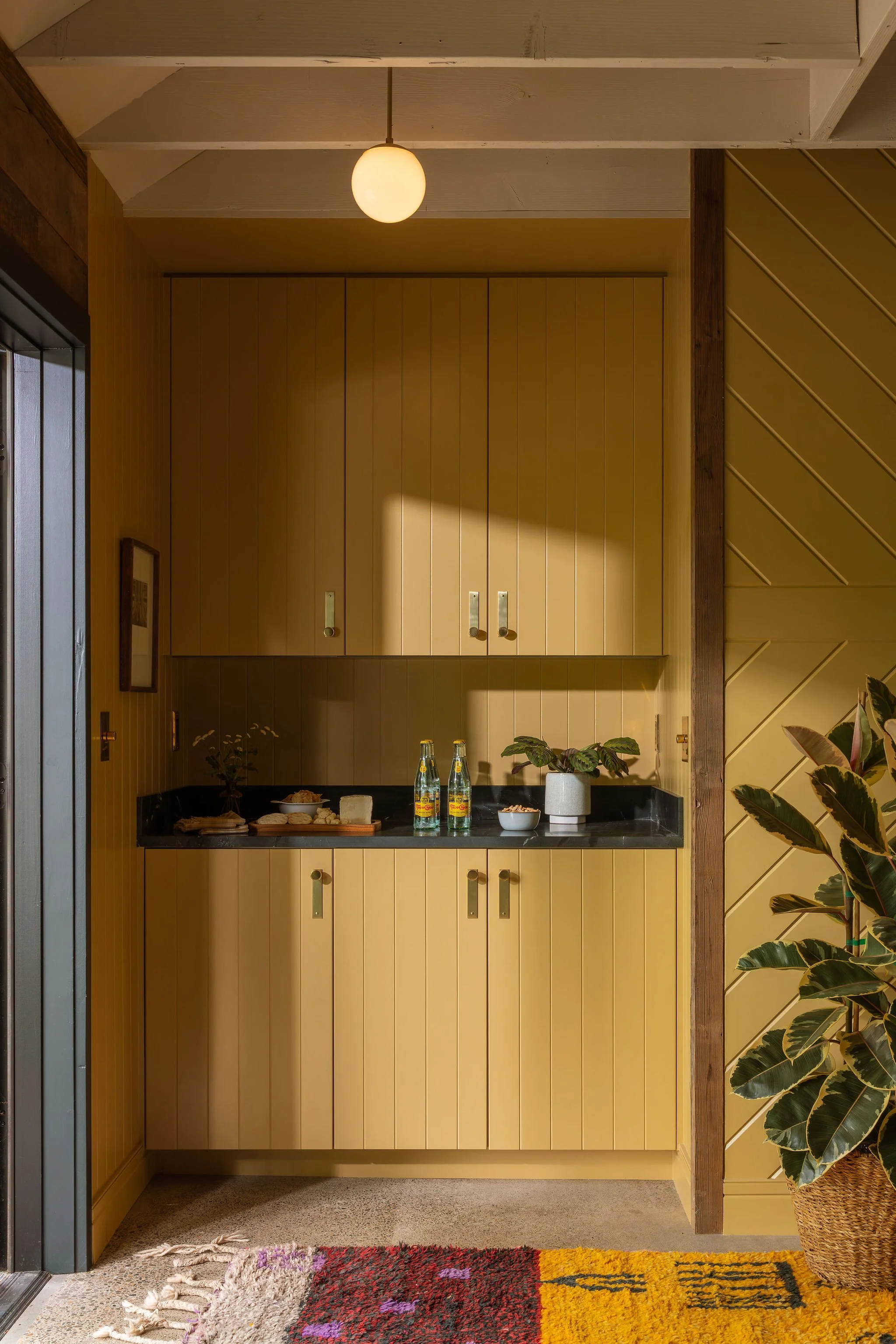 A yellow kitchen cabinet with a black countertop, decorated with bottles, plants, and food items, and flanked by a large plant in a wicker basket on the right side and a series of colorful rugs on the floor