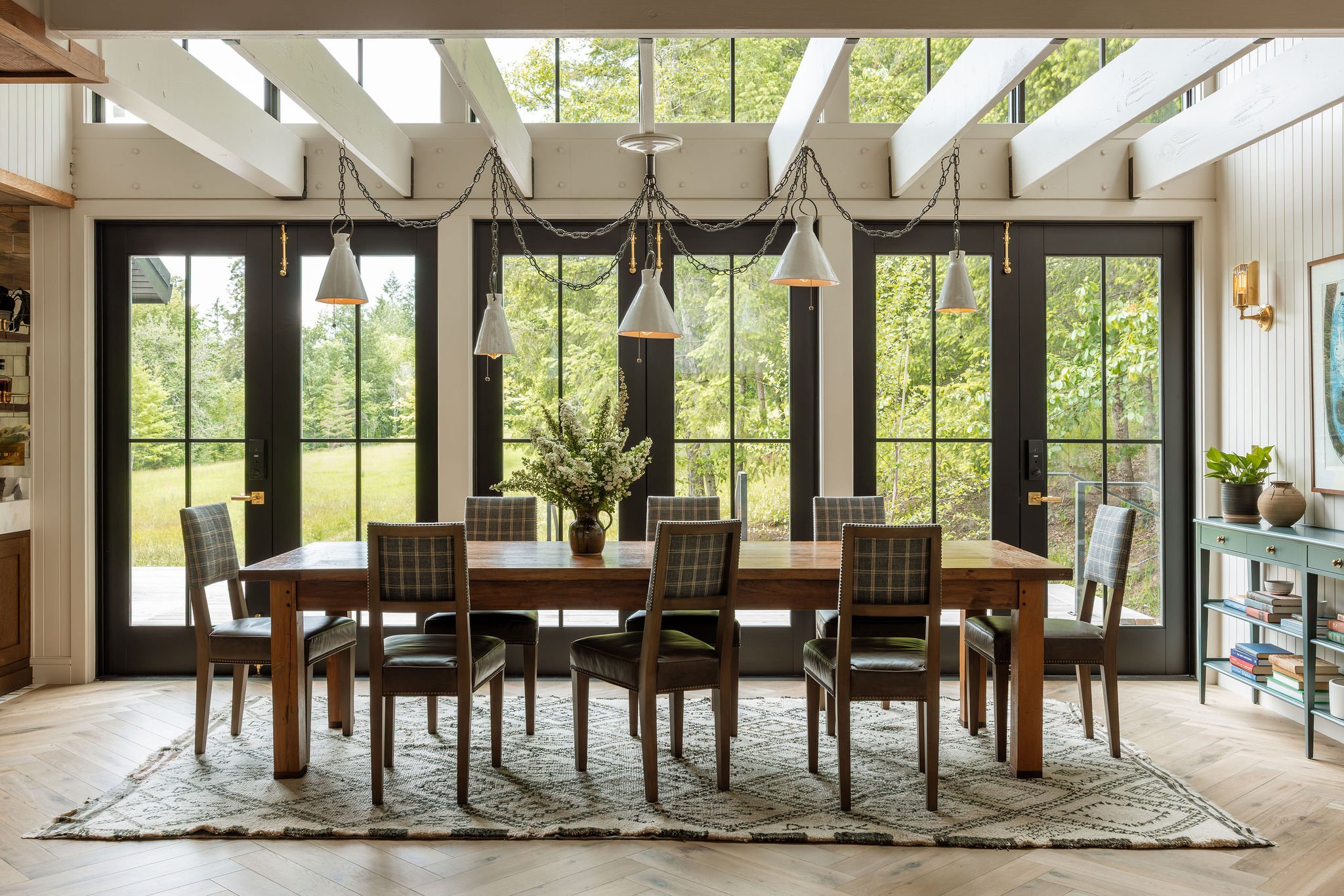 A dining room with a wooden table, plaid upholstered chairs, large black-framed glass doors, and a chandelier with layered metal shades hanging from chains. There is a large flower arrangement on the table, a patterned rug, and views of lush green tr