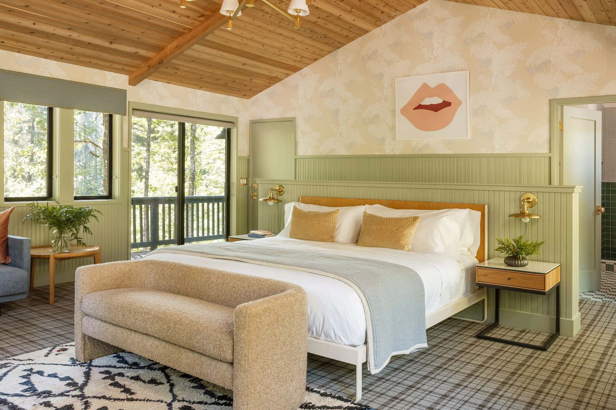 A cozy bedroom with a wooden vaulted ceiling, large windows showing trees outside, a bed with white sheets and beige pillows, a round beige ottoman at the foot, a small wooden side table with a green plant, and wall art depicting lips.