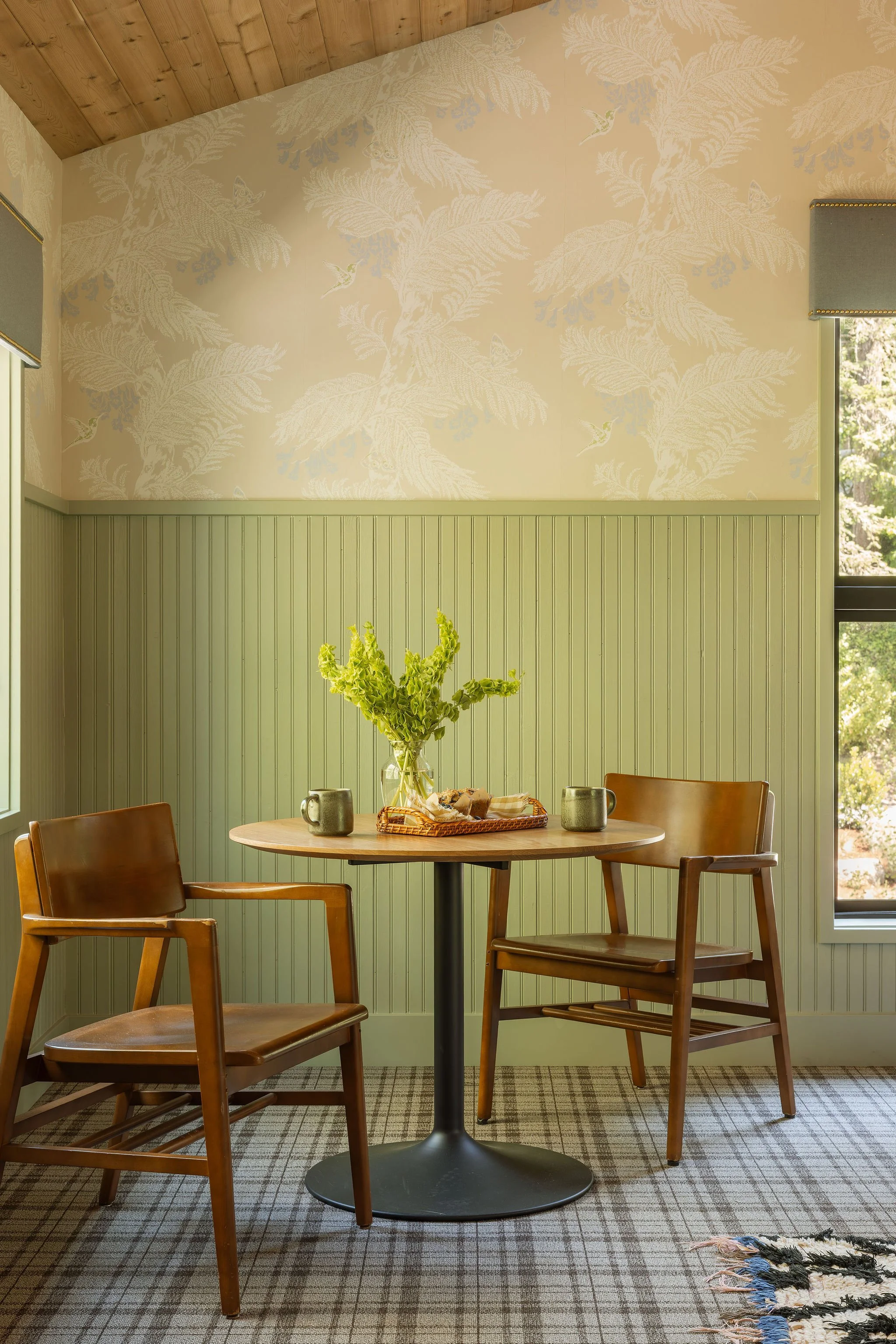A dining area with a wooden table and four wooden chairs. The table is set with a glass vase of green flowers, a plate of bread, two coffee mugs, and a small silver container. The walls are painted light green with wainscoting and beige wallpaper above featuring white, blue, and green botanical patterns with hummingbirds. There is a window with a small shade and a plaid carpet on the floor.