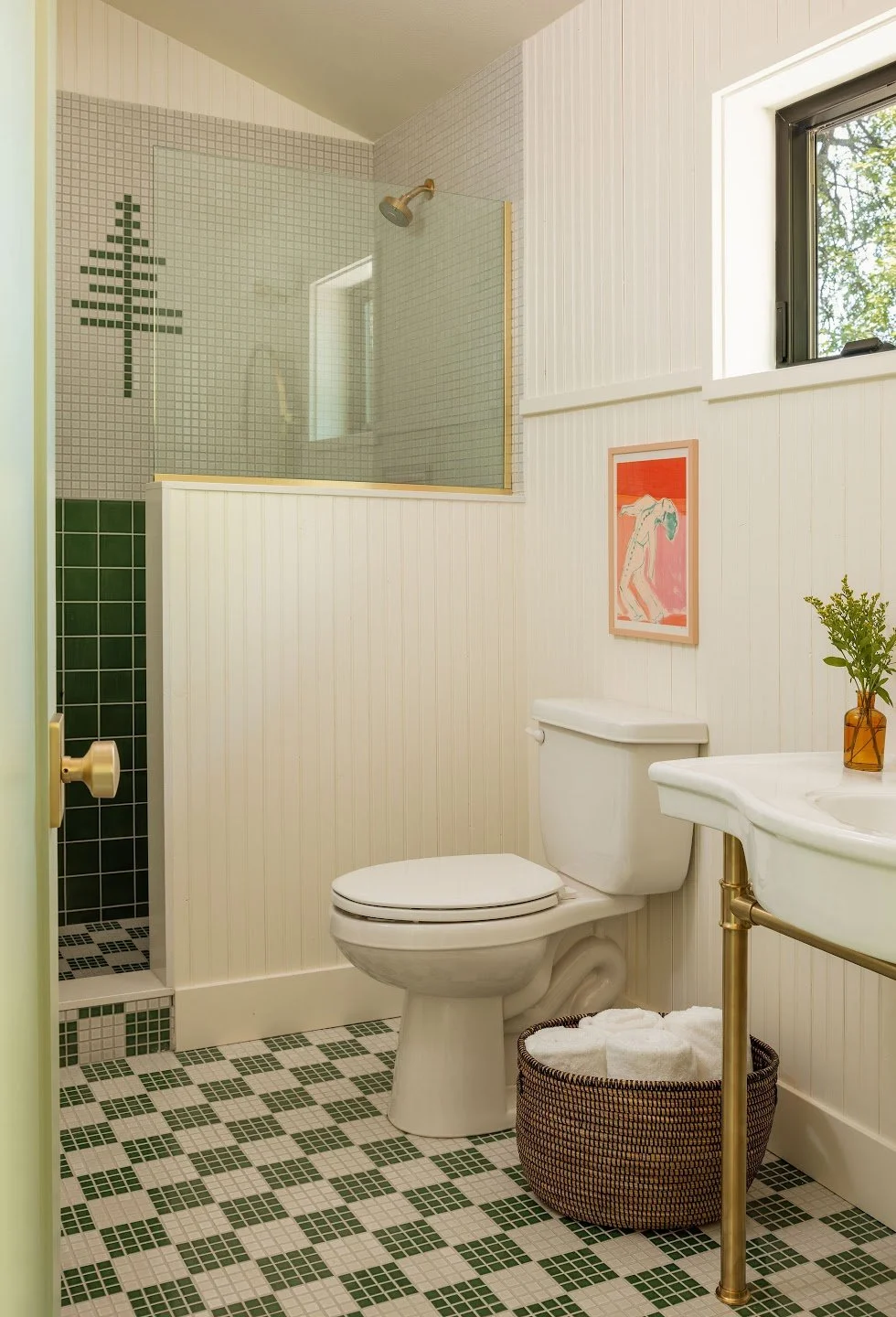 A small bathroom with a white toilet, a green tiled shower with a glass partition, a small sink with a gold-colored support, a framed artwork on the wall, a window with greenery outside, and a basket with rolled towels under the sink.