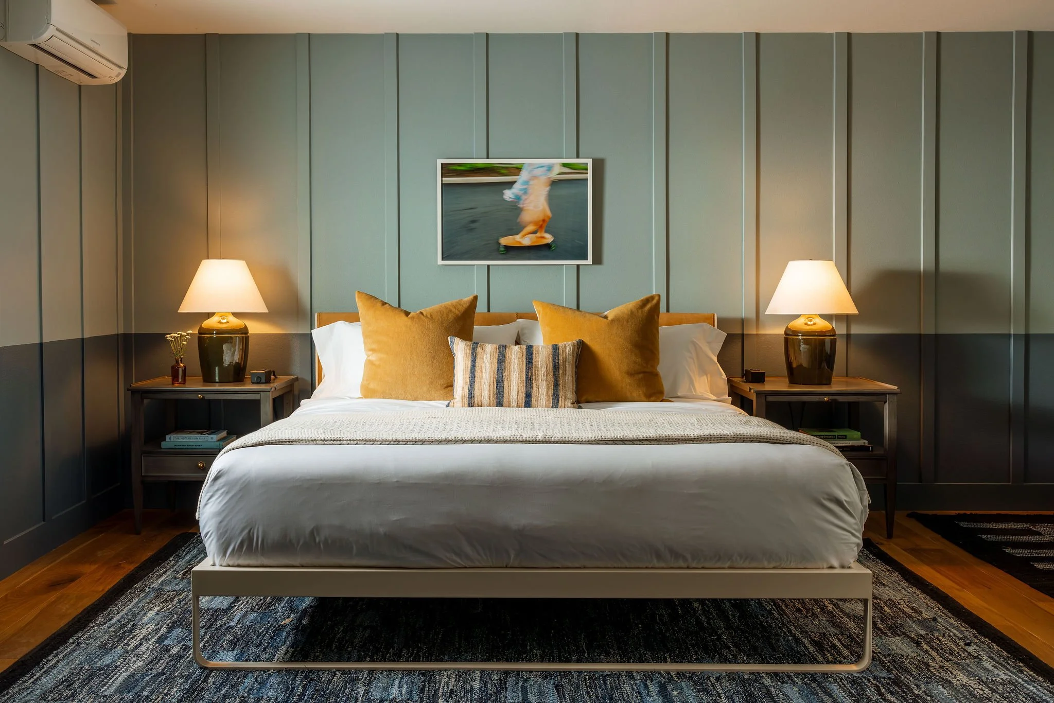 A modern bedroom with a large bed, tan pillows, and a striped throw pillow. Two matching bedside tables with lamps, a framed photo or artwork above the bed, and an air conditioning unit on the upper left wall. The room has blue wall panels, wooden floor, and a patterned area rug.
