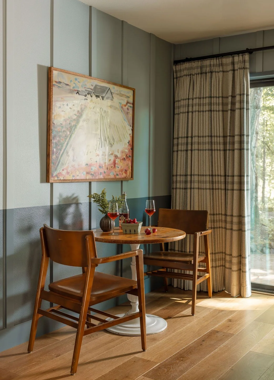 A cozy corner with a small wooden round table, two wooden chairs, a pitcher with foliage, two glasses of rosé wine, and a bowl of strawberries. There is a colorful landscape painting on the wall and a window with checkered curtains letting in natural light.