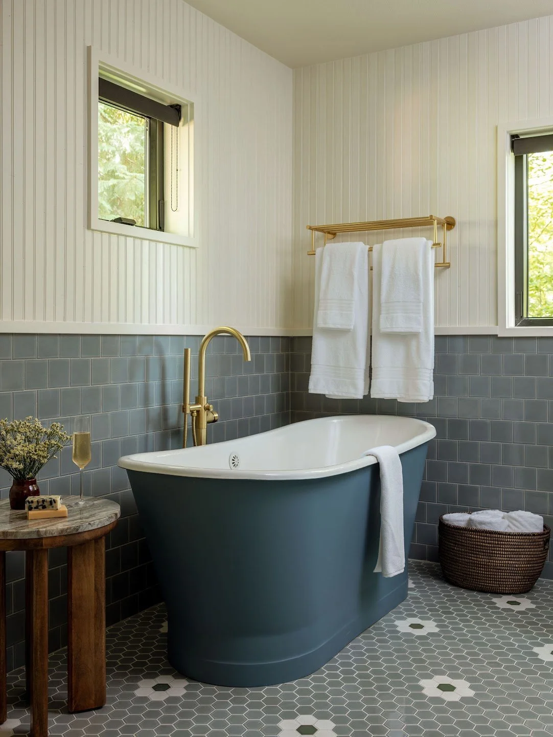 Bathroom with white beadboard walls, gray subway tiles, a dark blue bathtub with gold fixtures, two windows, white towels on a gold towel rack, a small round wooden table with decor, a wicker basket with towels, and a hexagon tile floor.