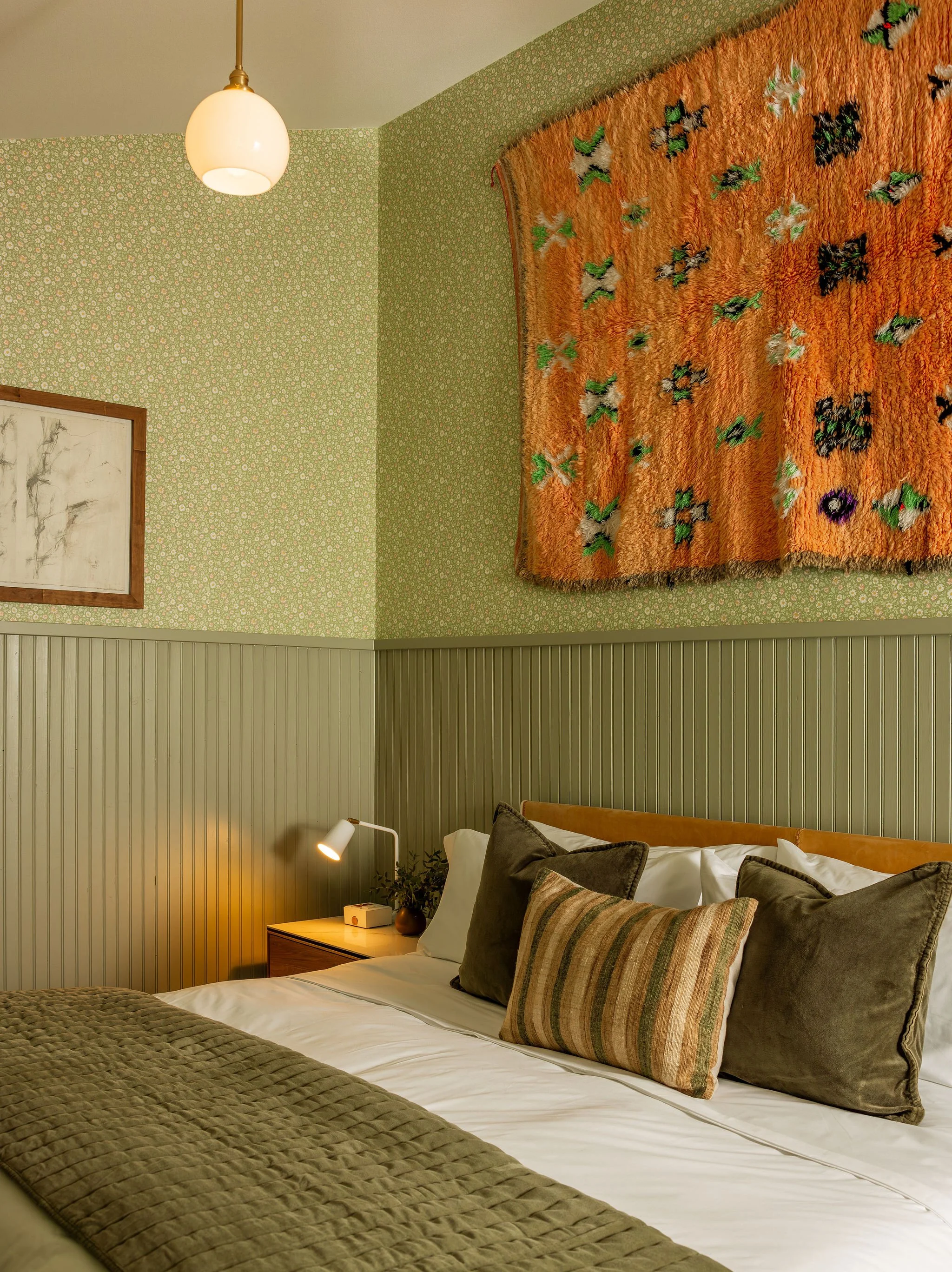 A bedroom with a bed, green beadboard wall, framed artwork, a bedside table with a lamp, and an orange textured wall hanging with embroidered patterns.