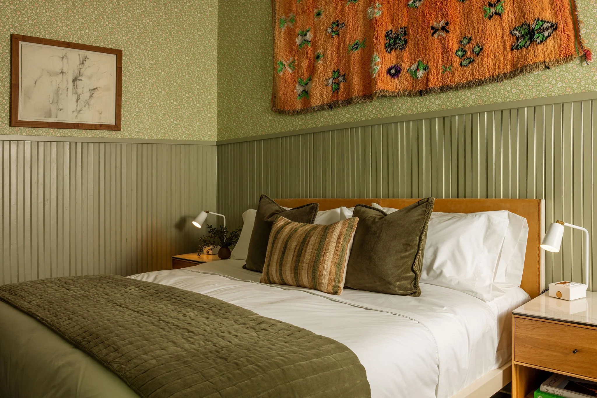 A neatly made bed with white sheets, a beige quilt at the foot, and multiple brown and green pillows, in a bedroom with green floral wallpaper, wooden wainscoting, a wooden nightstand with a desk lamp, a small potted plant, and a modern wall-mounted picture.