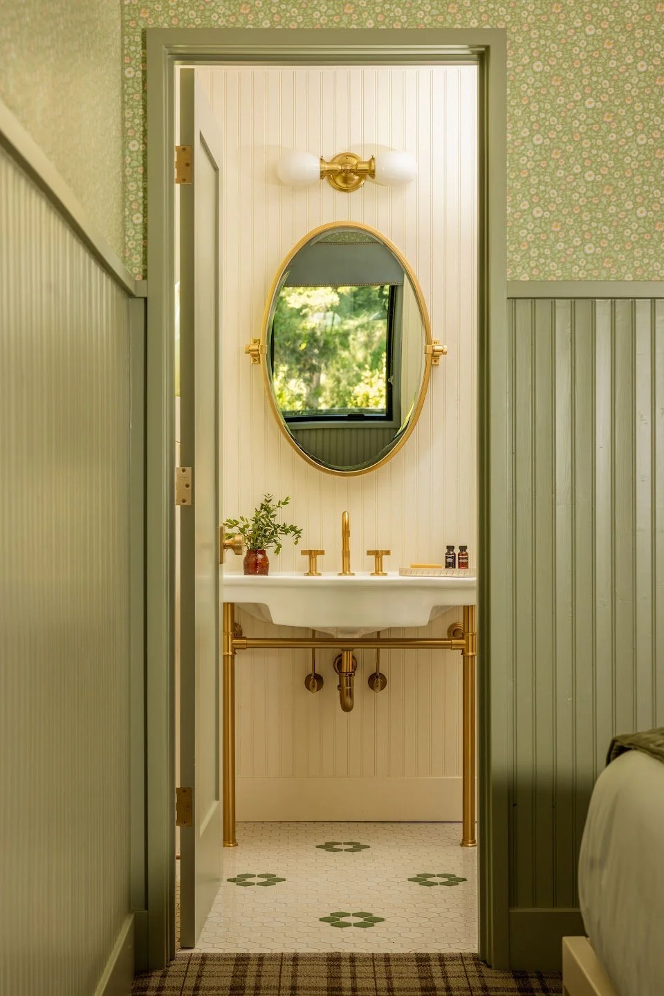 A small bathroom with a white sink and gold fixtures, an oval mirror, a small window reflected in the mirror, a potted plant, and wall-mounted light fixtures, with green wainscoting and floral wallpaper.