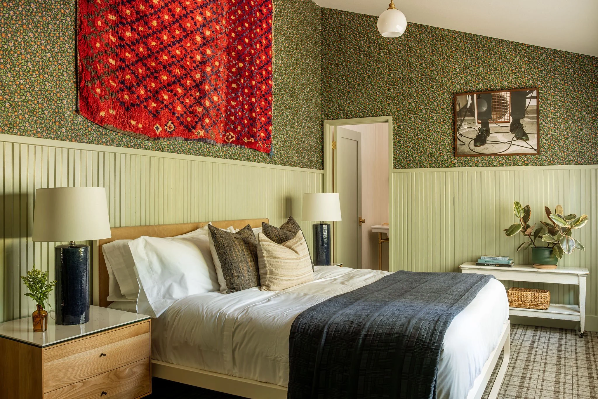 A bedroom with a bed, pillows, nightstand with a plant and a lamp, a mirror on the wall, and patterned wallpaper.