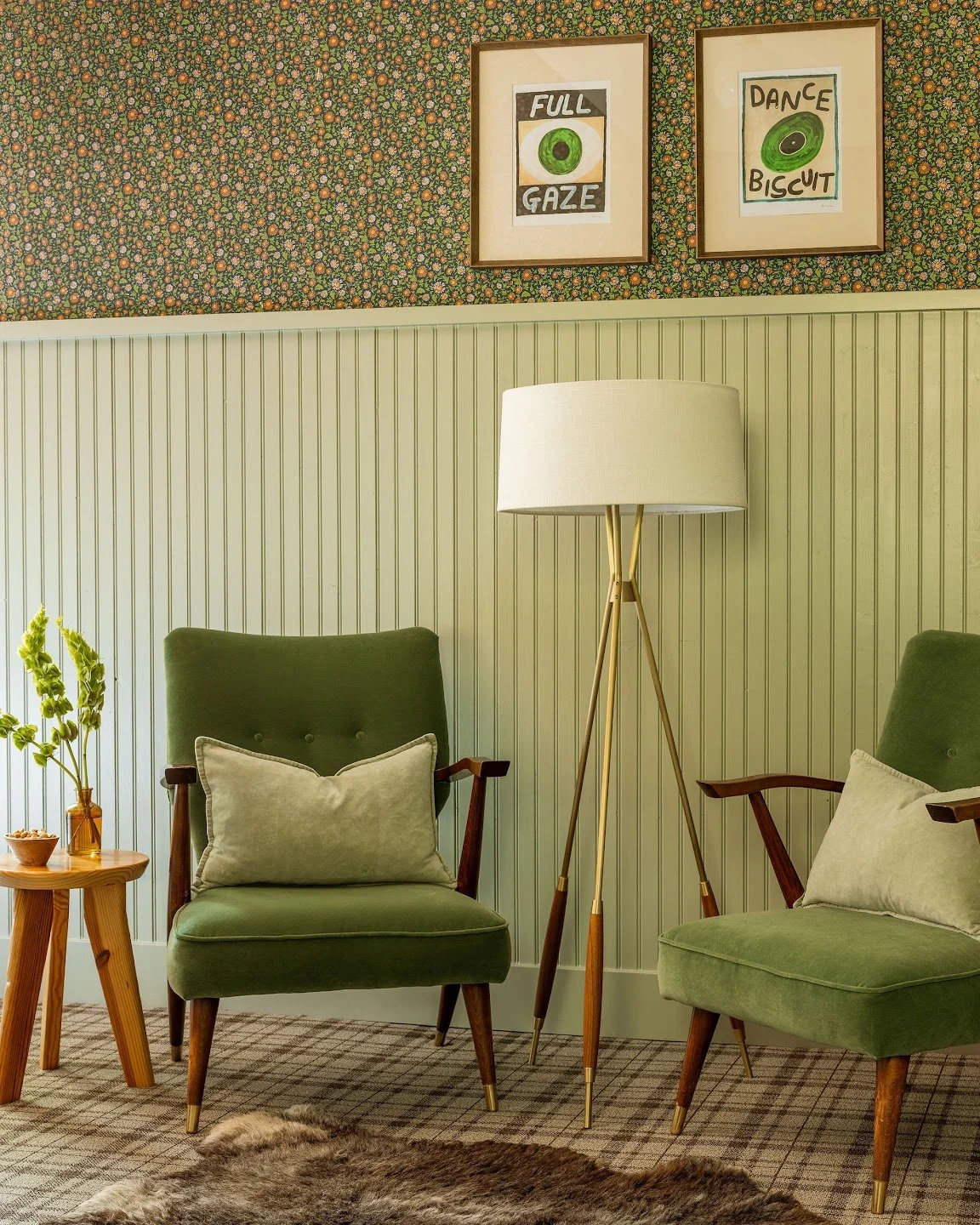 A cozy living room corner with two green armchairs, a wooden side table with a plant, a tall floor lamp, and framed wall art.
