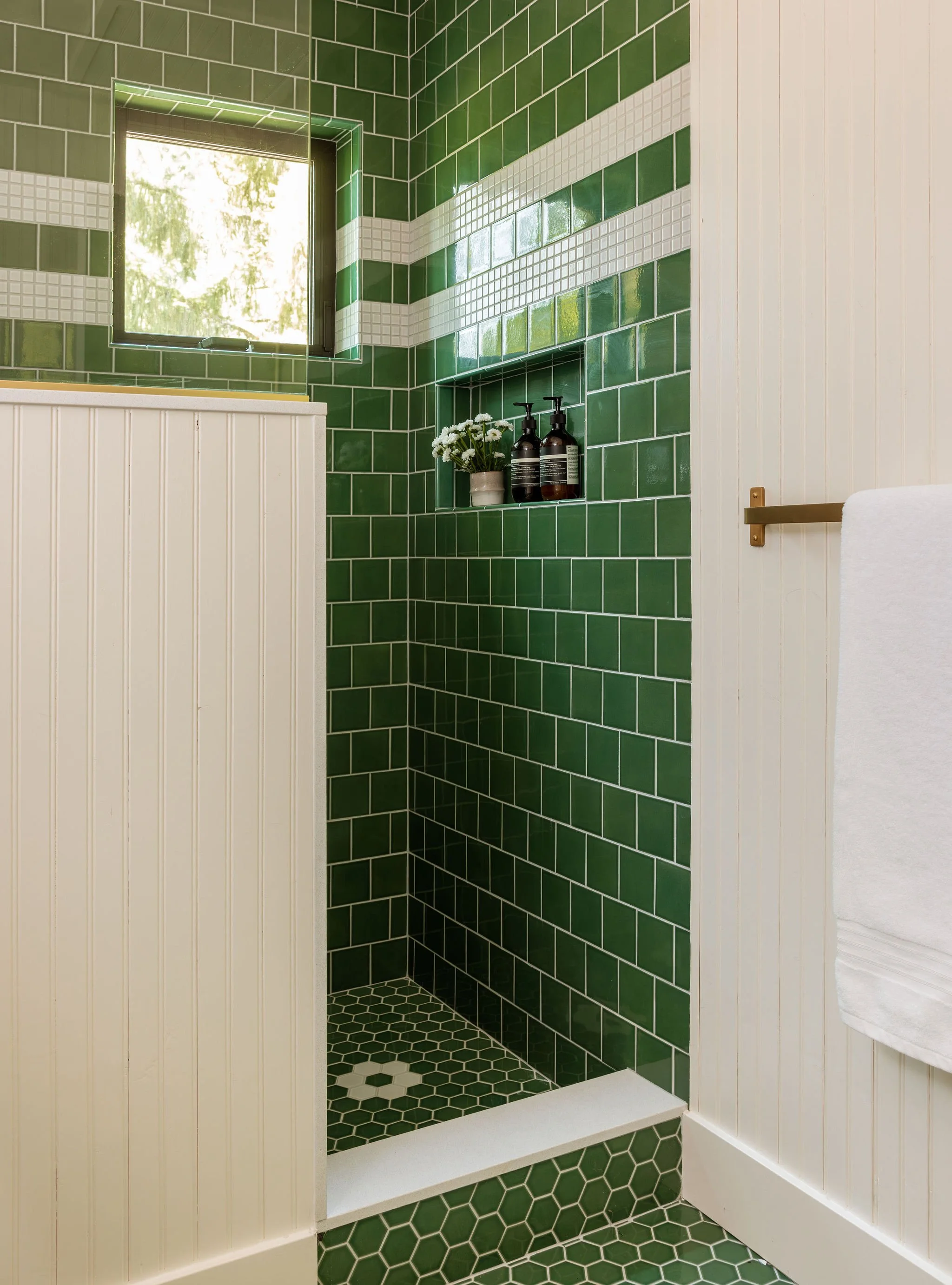 Bathroom with green and white tiled shower area, a small window with natural light, a built-in shelf holding a potted plant and soap dispensers, white beadboard walls, and a white towel hanging on a towel holder.