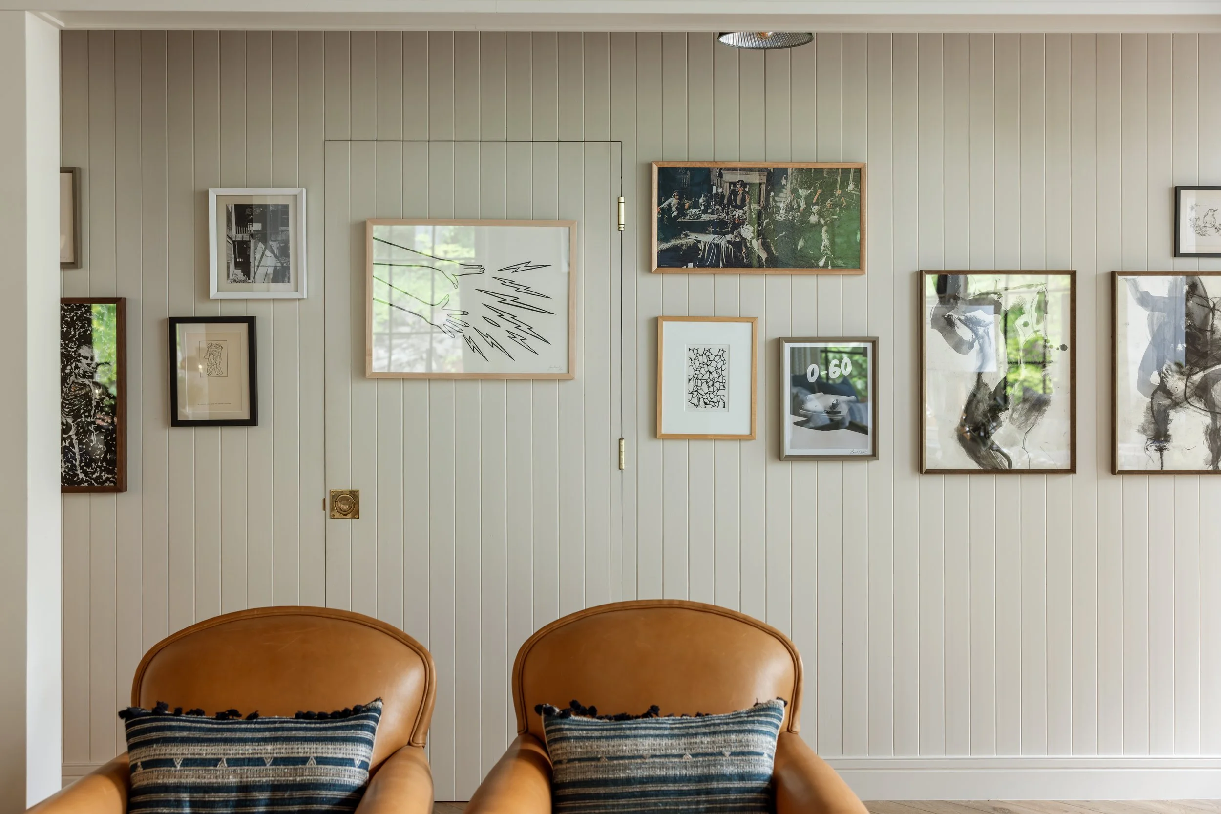 Living room with light-colored paneled wall decorated with various framed artwork, two brown leather chairs with striped throw pillows, and partially visible ceiling light fixture.