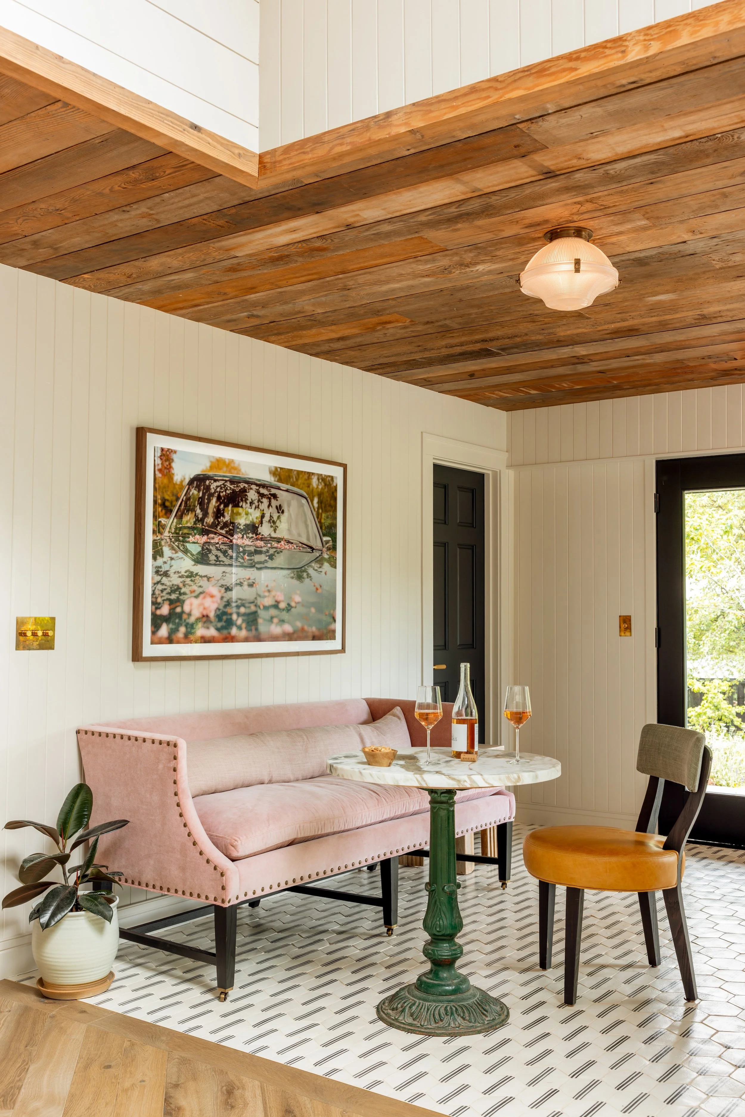 A cozy living room with a pink velvet sofa, a round marble-topped table with drinks, a black and a tan chair, a framed picture of a car with pink flowers, a white wall with paneling, a wooden ceiling, and a patterned tile floor.