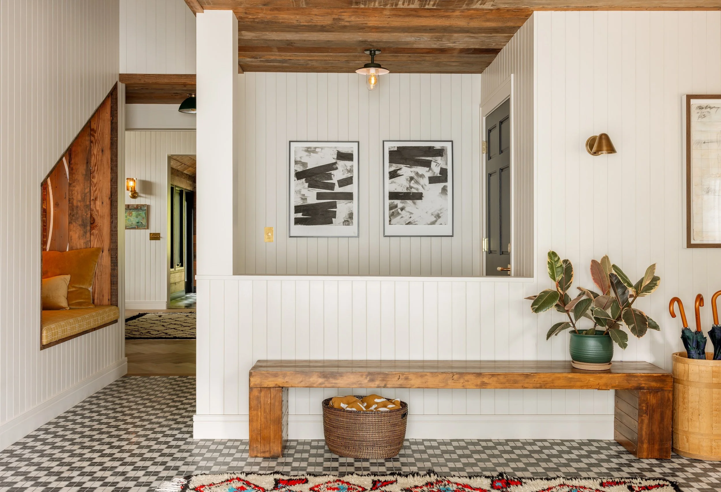A cozy, modern entryway with white beadboard walls, a wooden ceiling, and Asian-inspired decor. There is a black-and-white abstract art piece on the wall, a built-in window seat with a yellow cushion, and a large green potted plant. The floor feature