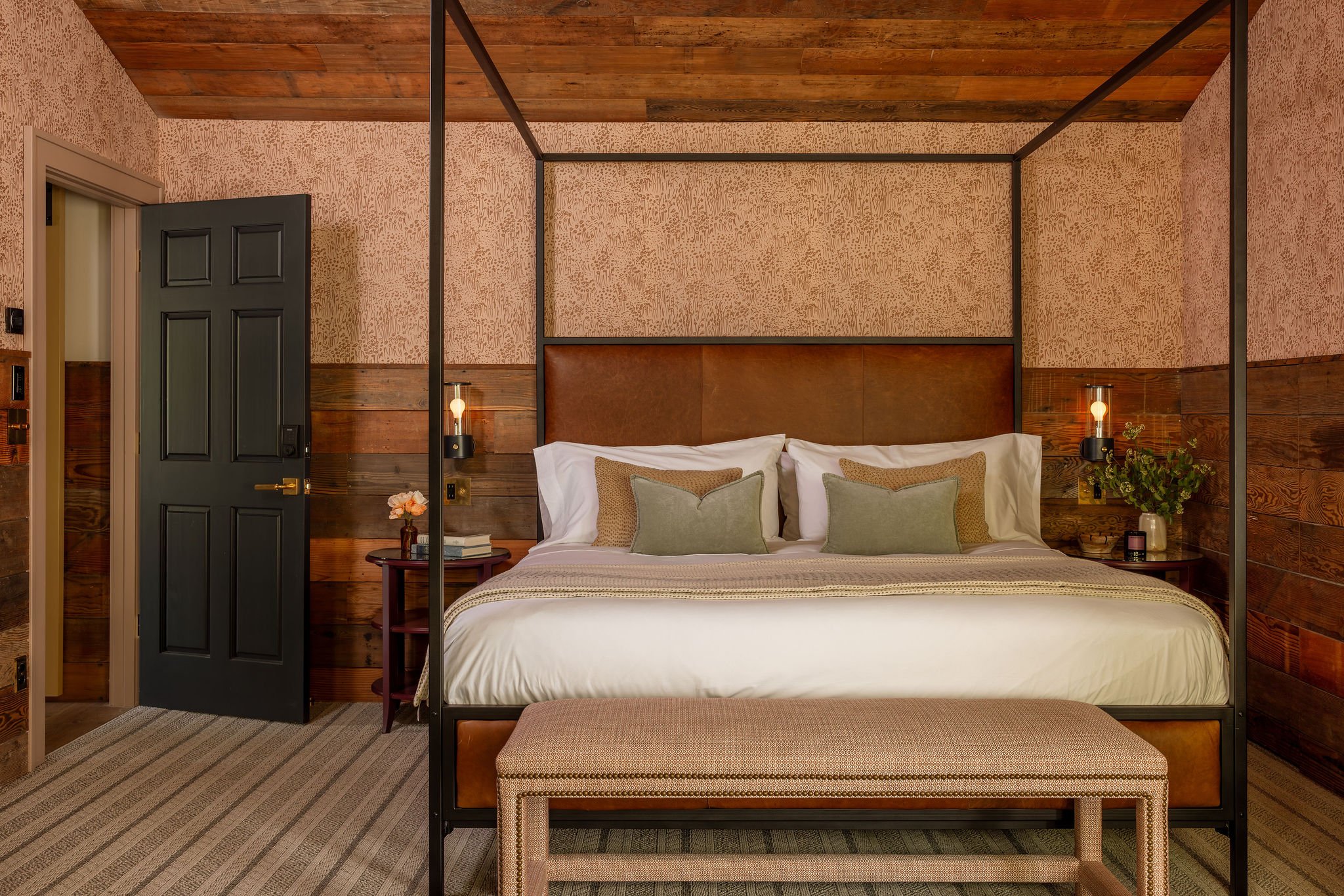 A bedroom with a bed that has a wood headboard, four pillows, and four lamps on the wall behind it. There is a black door to the left and a small round table with a flower and books. The room has wooden walls and ceiling with a patterned carpet on the floor.