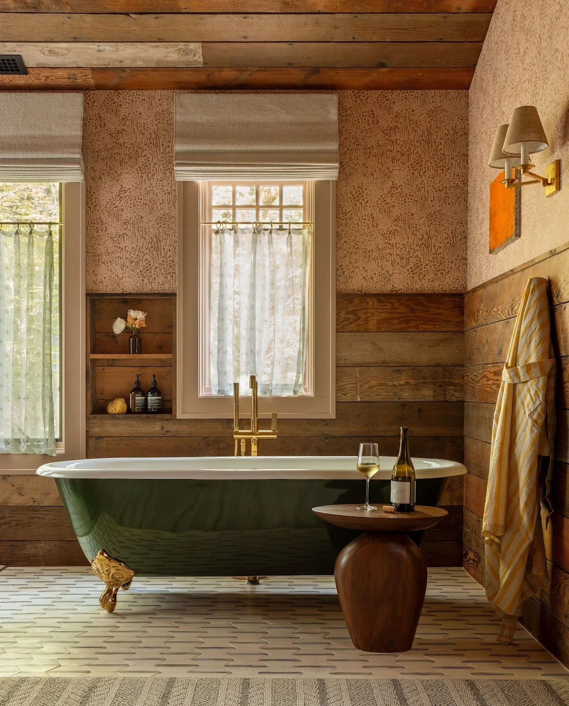 A cozy bathroom with a green clawfoot bathtub, a small wooden side table with a glass of white wine and a bottle, and wooden wall paneling. There are two windows with curtains, warm lighting, and a towel hanging on the wall.