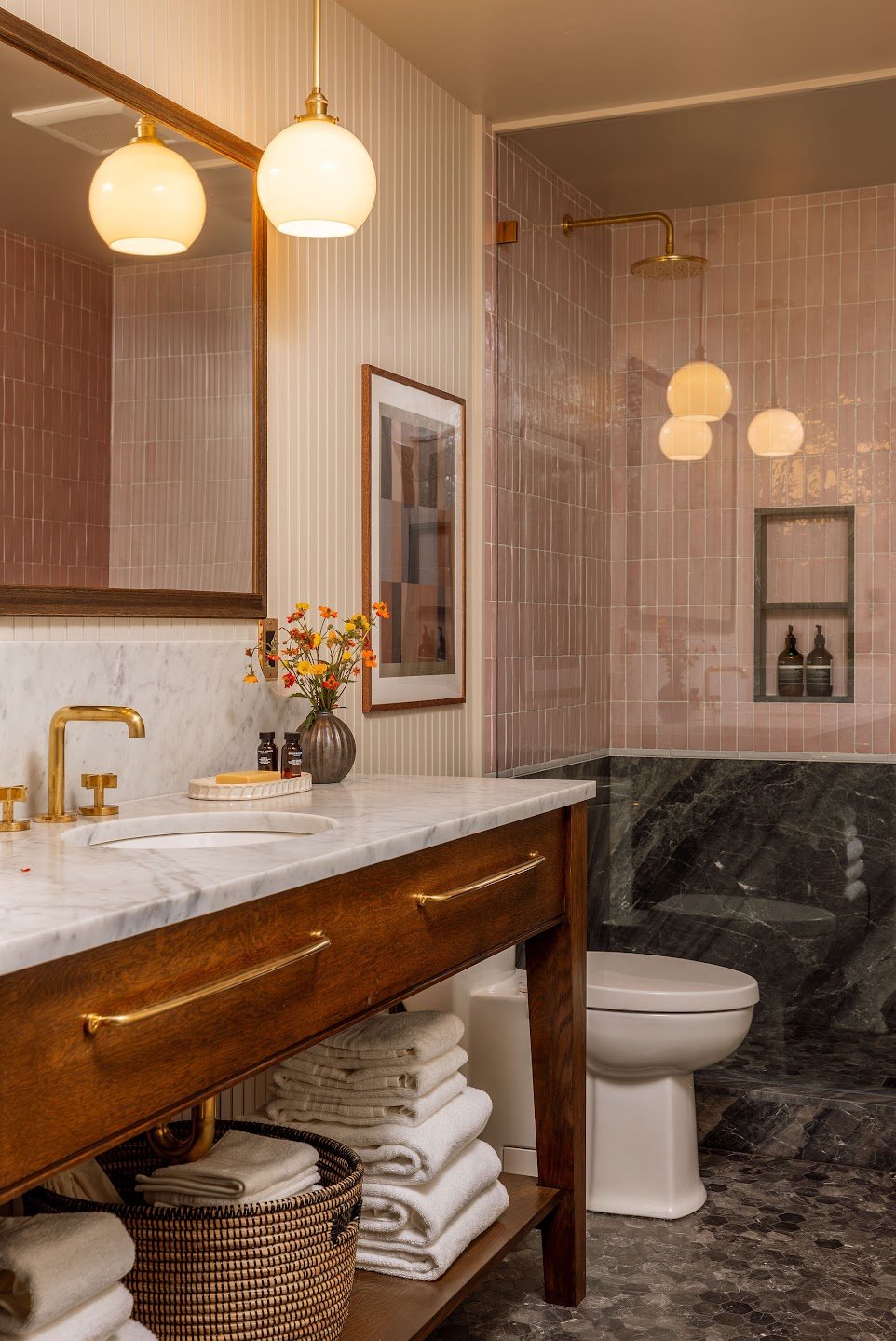 A modern bathroom with a marble countertop and brass fixtures, pink tiled shower area, and wooden vanity with hanging towel storage.