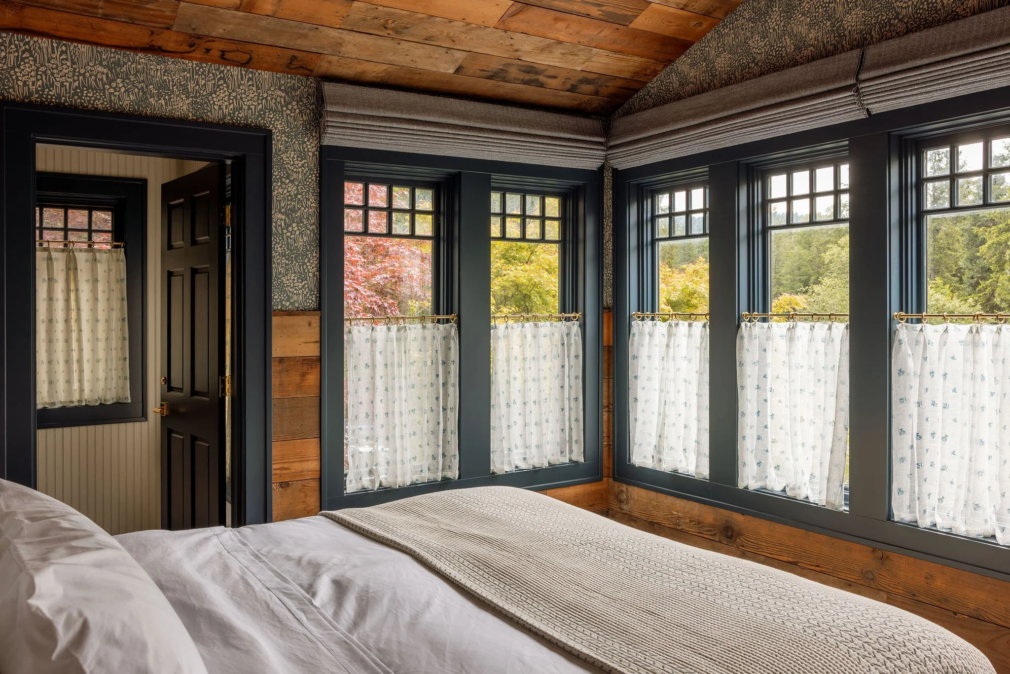 Room with large windows showing trees outside and white curtains hanging from a gold curtain rod, with a bed in the foreground and wooden walls and ceiling.