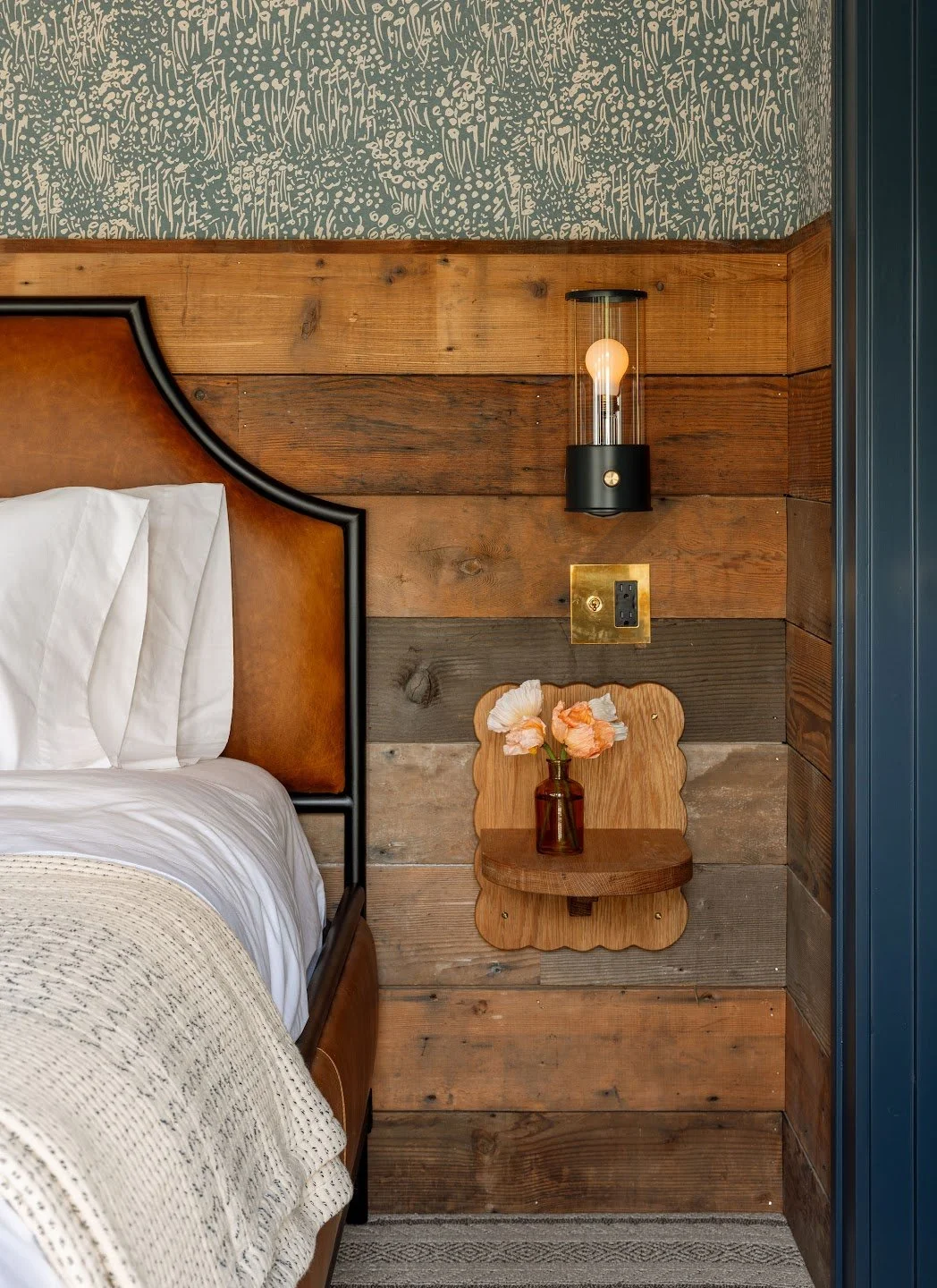 Close-up of a cozy bedroom corner with a wooden headboard, patterned wallpaper, modern wall sconce, and a small wooden shelf with a vase of pink flowers.