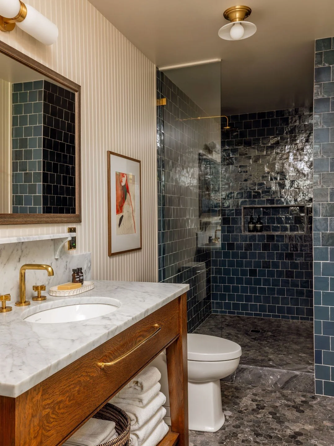 Modern bathroom with a stone shower, marble vanity with gold fixtures, and framed artwork on cream beadboard wall.