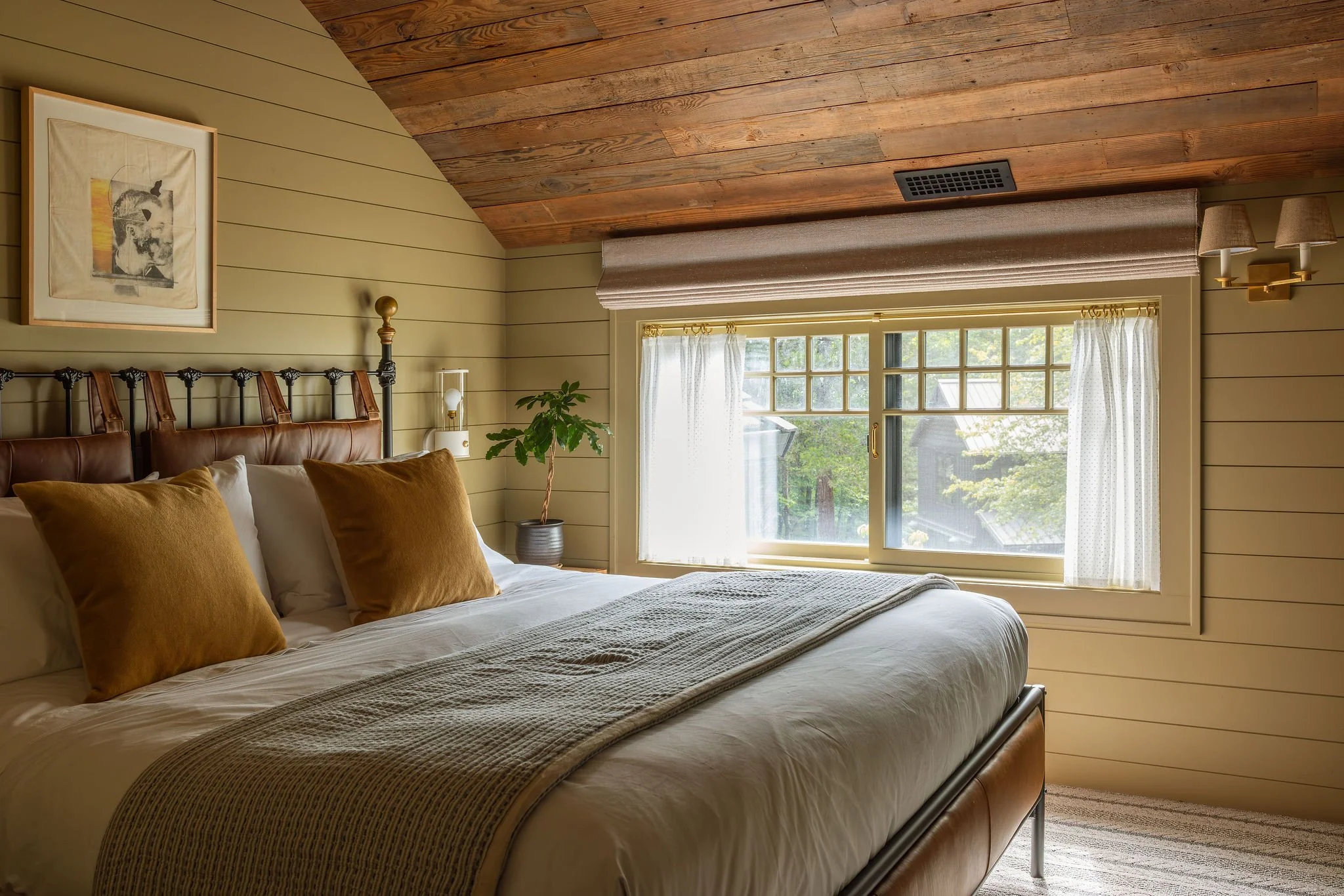 A cozy bedroom with a bed, brown pillows, and a beige and cream color scheme. The room has a large window with white curtains, a wooden ceiling, and wall panels, with a small potted plant on a side table.