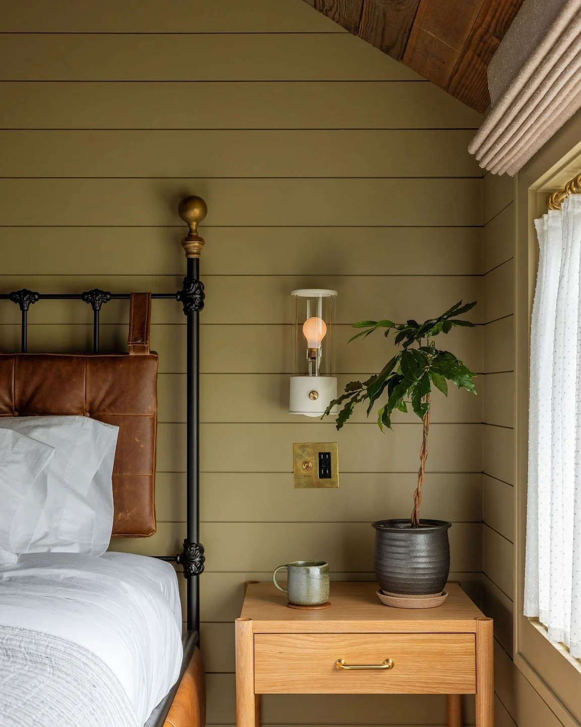A cozy bedroom features a bed with a leather headboard, a nightstand, a potted plant, a wall-mounted lamp, and light-colored shiplap walls near a window with curtains.